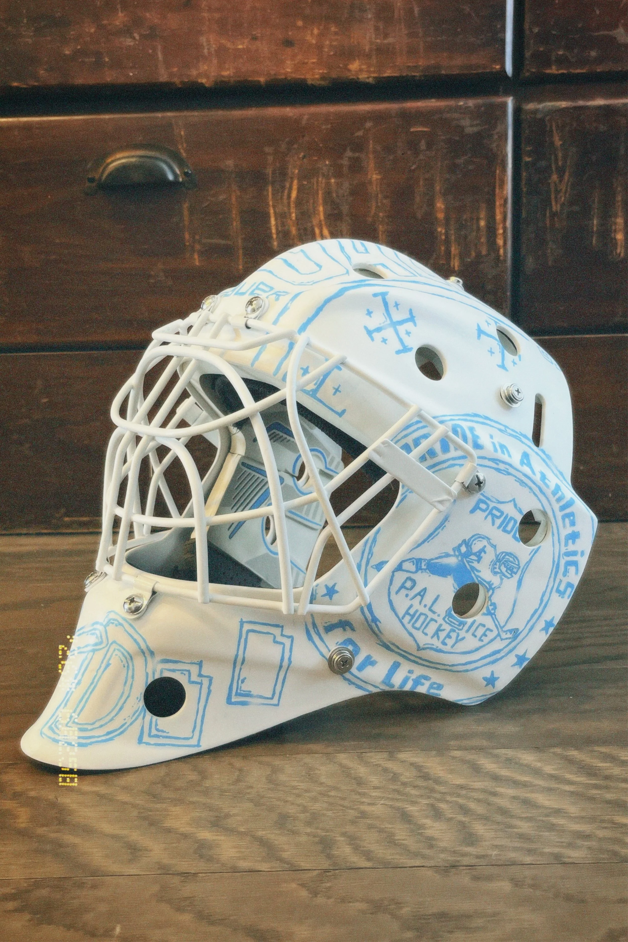 A hockey goalie mask with a white and blue design, featuring the words "P.A.L. Hockey" and various patterns, resting on a wooden surface.