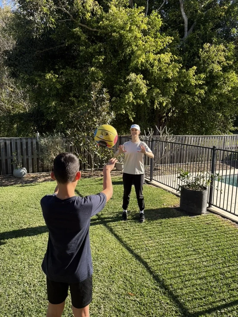 A young boy and a woman playing with a yellow and blue beach ball in a backyard with green grass, trees, and a fenced pool area.
