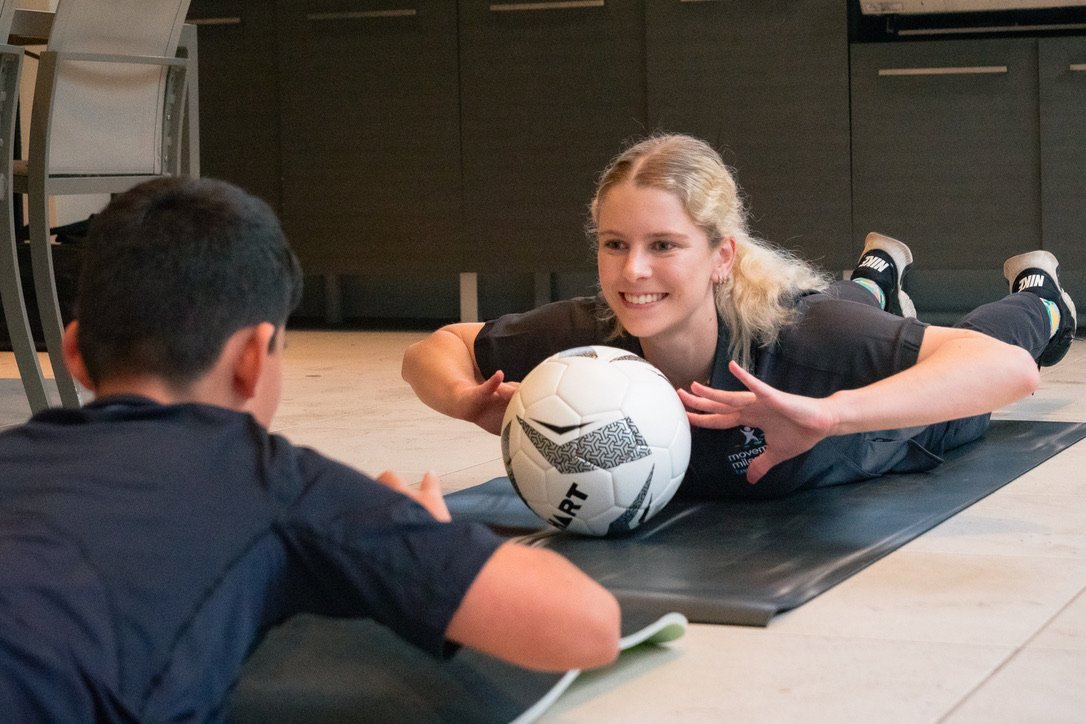 A woman with blonde hair doing a martial arts move on a black mat, with a soccer ball under her chin, facing a young boy lying on the mat across from her.