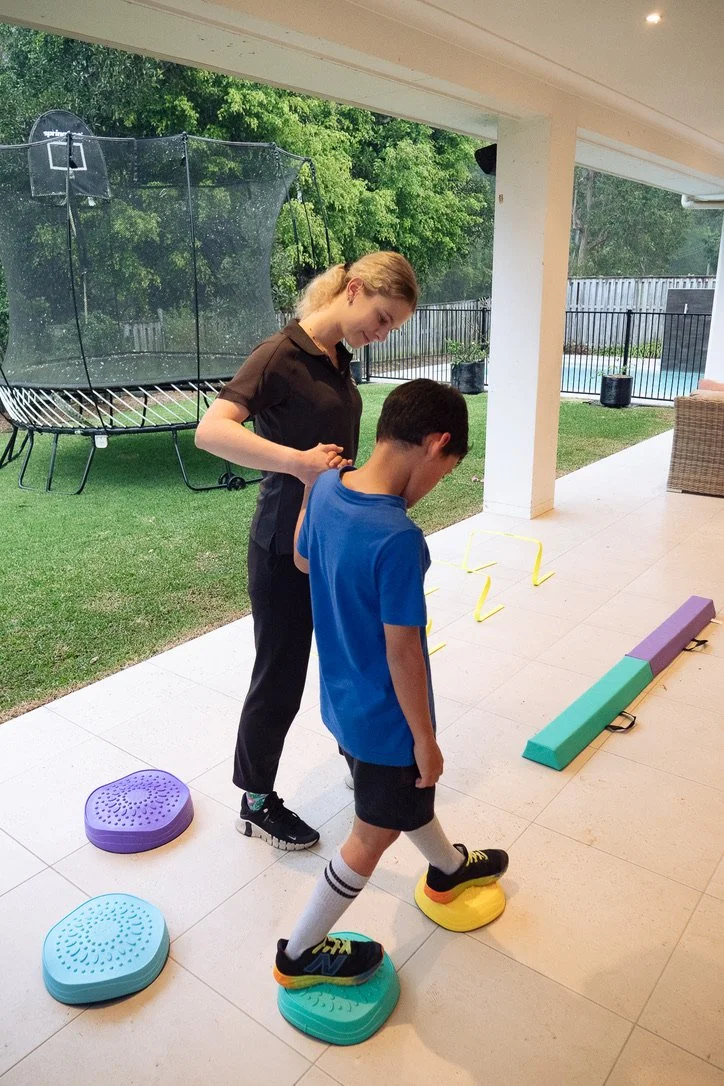 A woman helping a boy with a physical therapy exercise outside on a porch, with agility training equipment in the background.