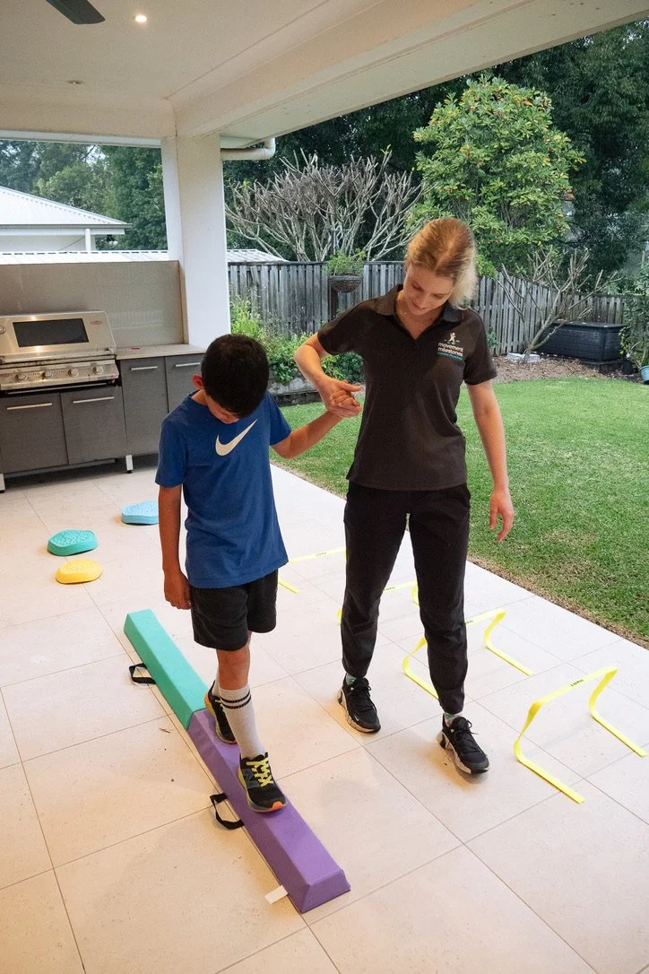 A young boy practices walking on a balance beam with assistance from a woman, in an outdoor exercise area with agility hurdles and training equipment.