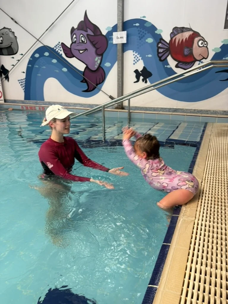 A young girl in a pink swimsuit with colorful patterns is sitting at the edge of a swimming pool, splashing water towards a woman in a maroon long-sleeve shirt and a white hat, who is standing in the pool. The background features a mural of cartoon fish and a purple mermaid.