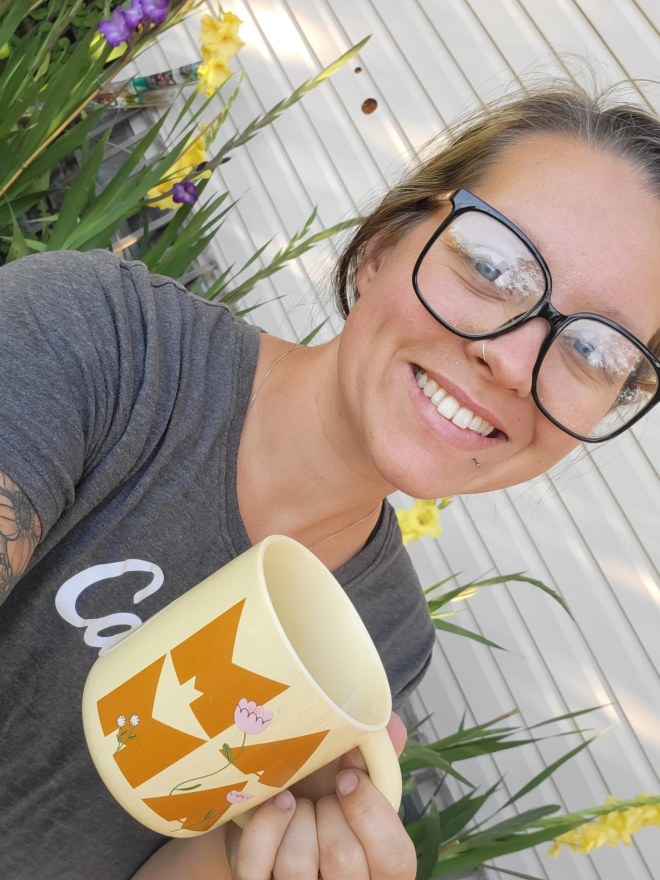 A woman smiling, wearing glasses and a gray shirt, holding a pumpkin-shaped mug with a jack-o'-lantern face and floral decorations, outdoors near white siding and yellow and purple flowers.