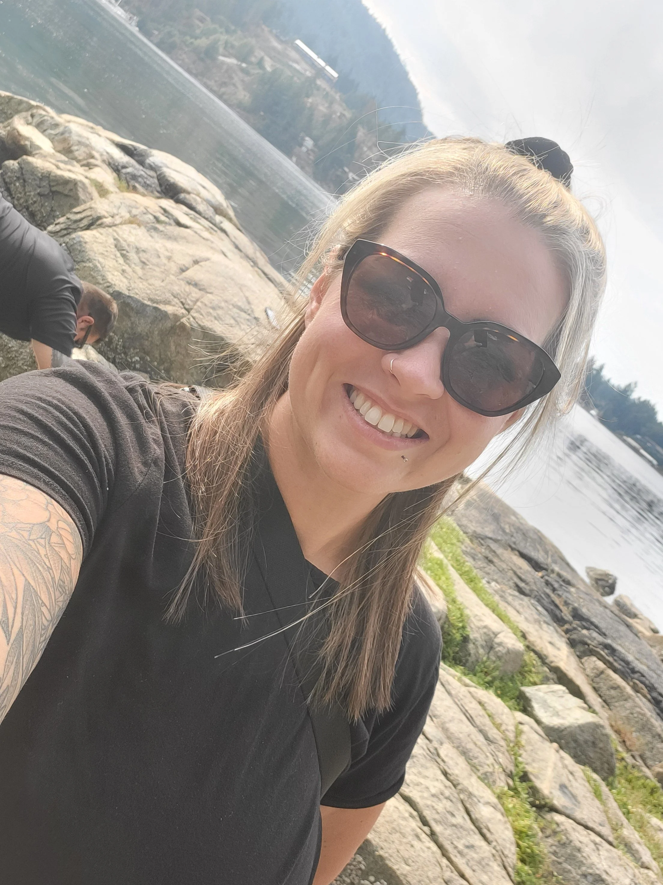 A woman with blonde hair, sunglasses, and a black shirt taking a selfie by a rocky shoreline with water and a mountain in the background.