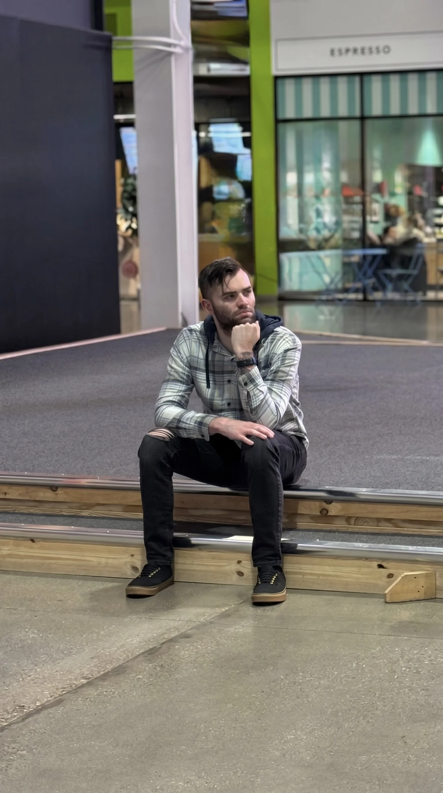 A young man with a beard and short brown hair, wearing a plaid shirt, black pants with ripped knees, and black sneakers, sitting on a wooden platform with his chin resting on his hand, inside a shopping mall or similar indoor space.