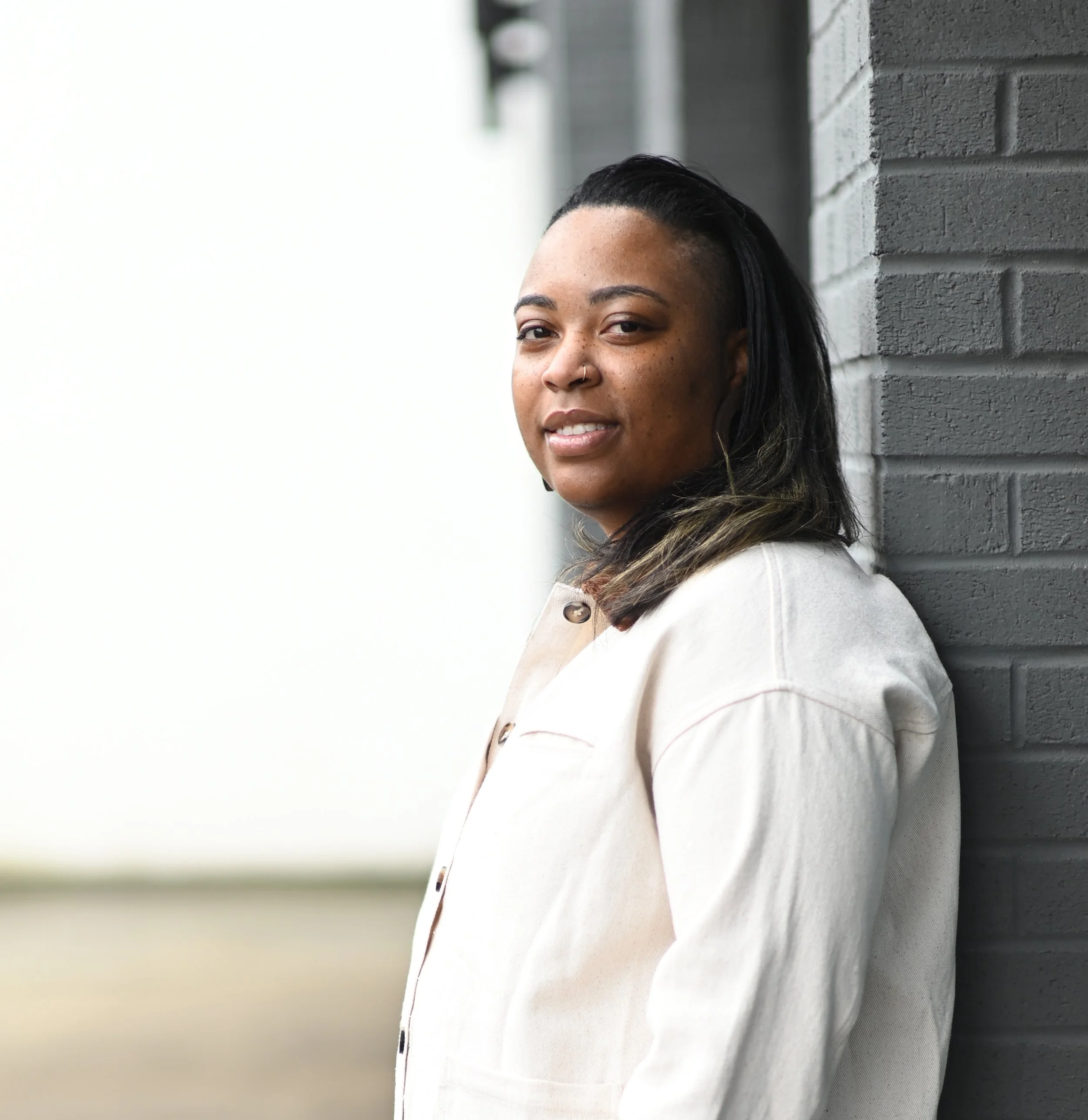 Woman with dark hair and earrings leaning against a brick wall, looking at the camera.