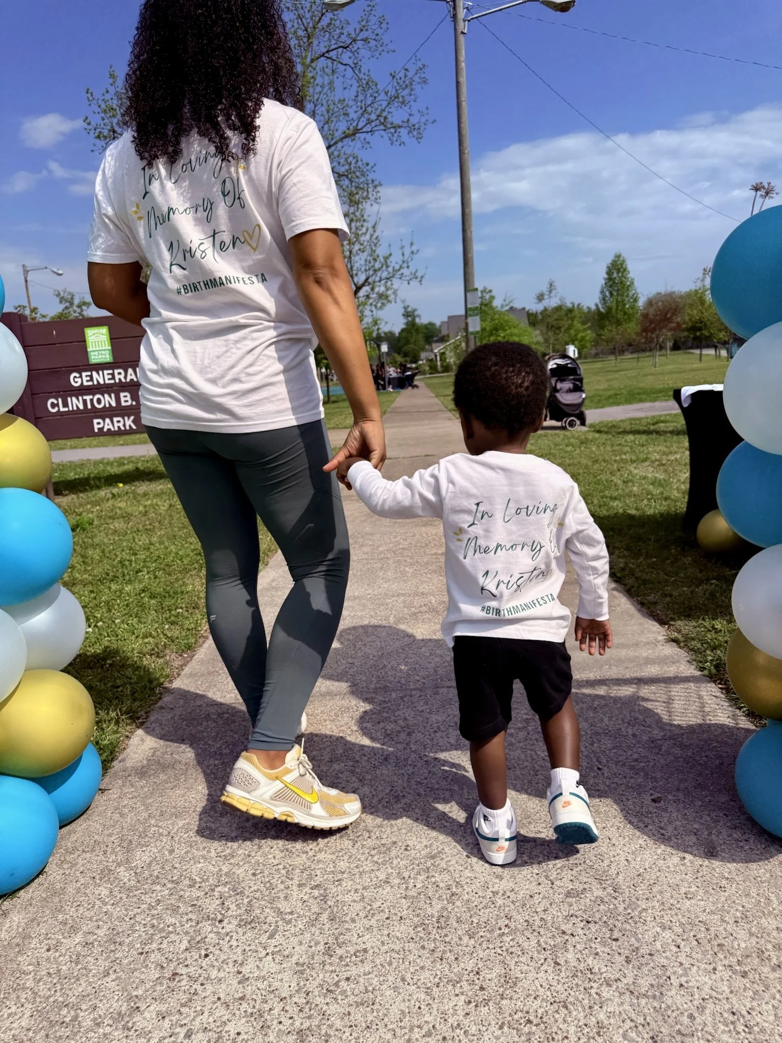 A woman and a young boy walk hand in hand through a park decorated with blue, white, and yellow balloons. The woman is wearing a white T-shirt with text that reads 'In Loving Memory Of Kristen #BIRTHMANIFESTA' and gray leggings. The boy is dressed in