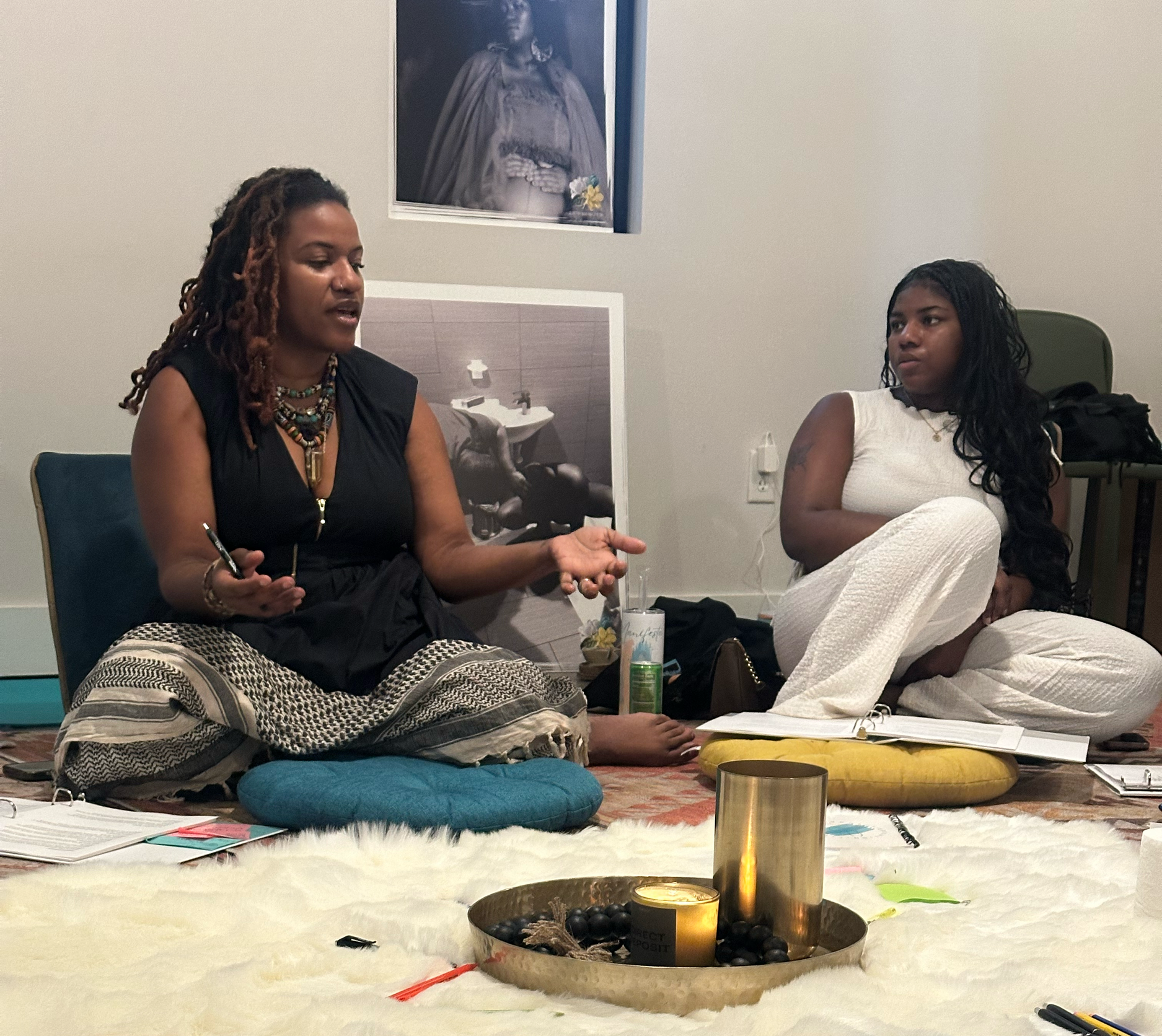 A woman with red dreadlocks, wearing a black top and colorful beaded necklaces, is sitting at a table with various office supplies and notebooks. She is gesturing with her hands while talking to someone. The table is covered with a white furry tablec