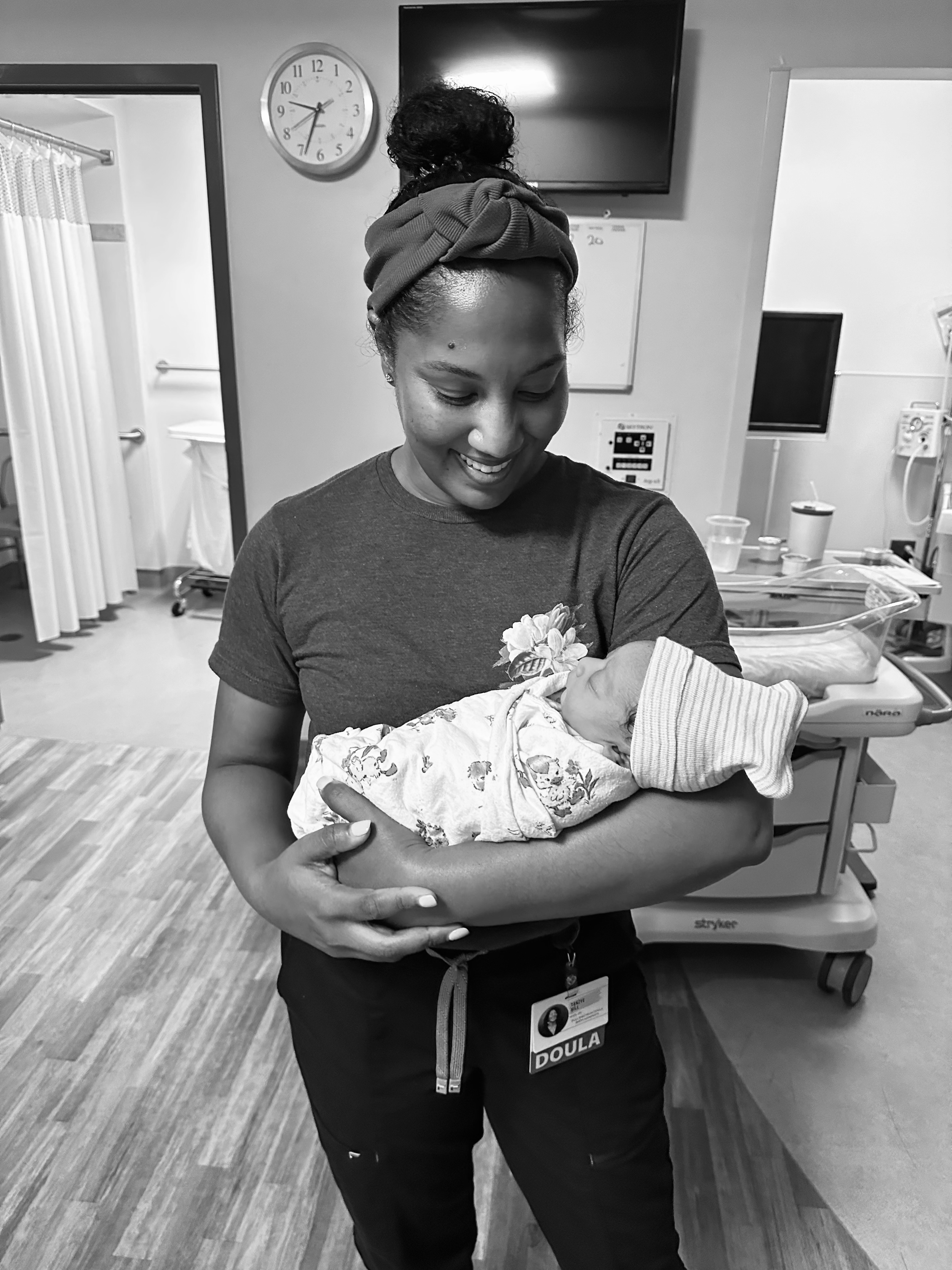 A woman in hospital scrubs holding a newborn baby wrapped in a blanket, standing in a hospital room with medical equipment.