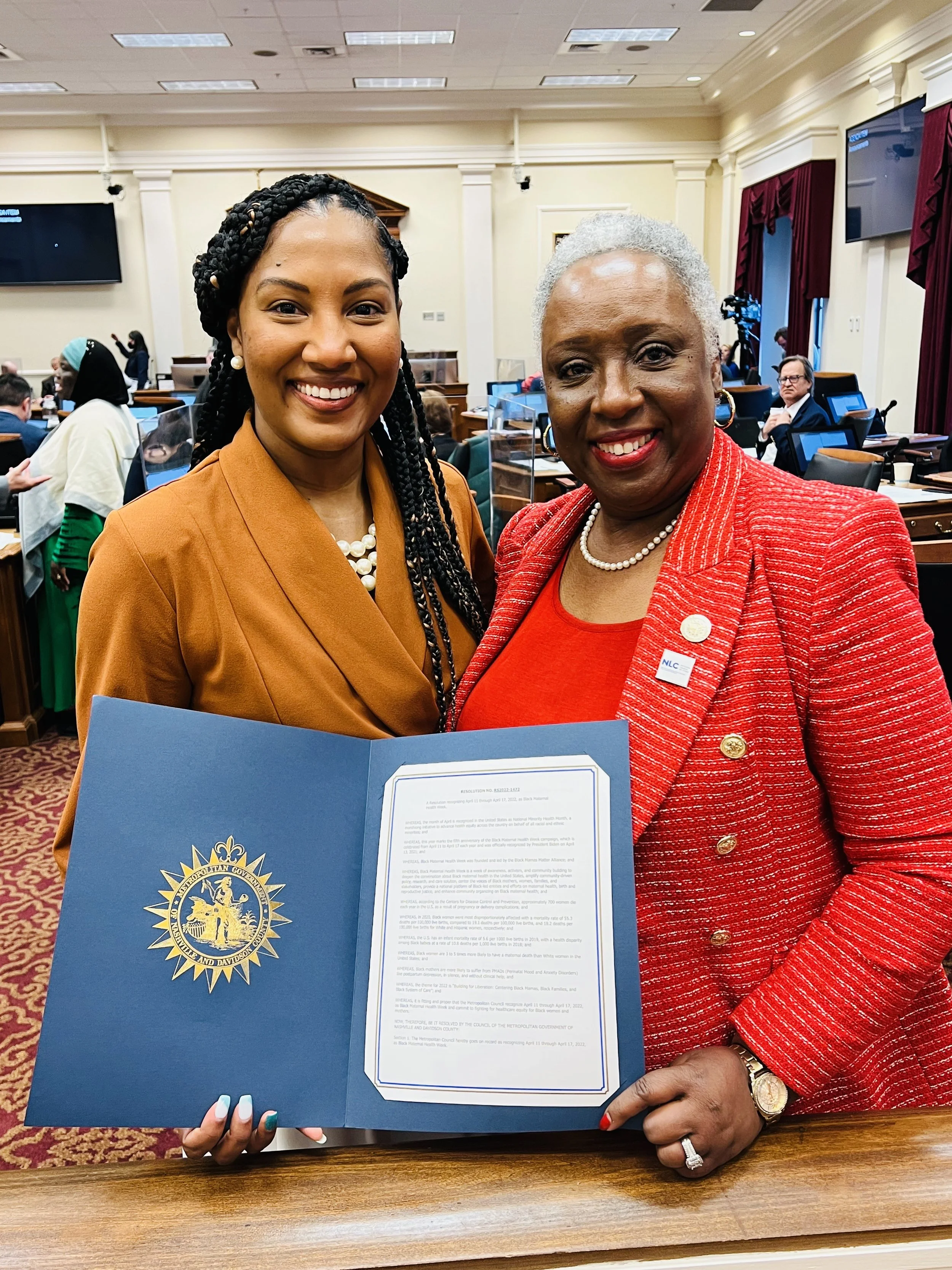 Two women standing in a meeting room, smiling. One woman is holding a document with an official emblem. The woman on the left is wearing a tan blazer with a pearl necklace, and the woman on the right is wearing a red blazer with a pearl necklace and 