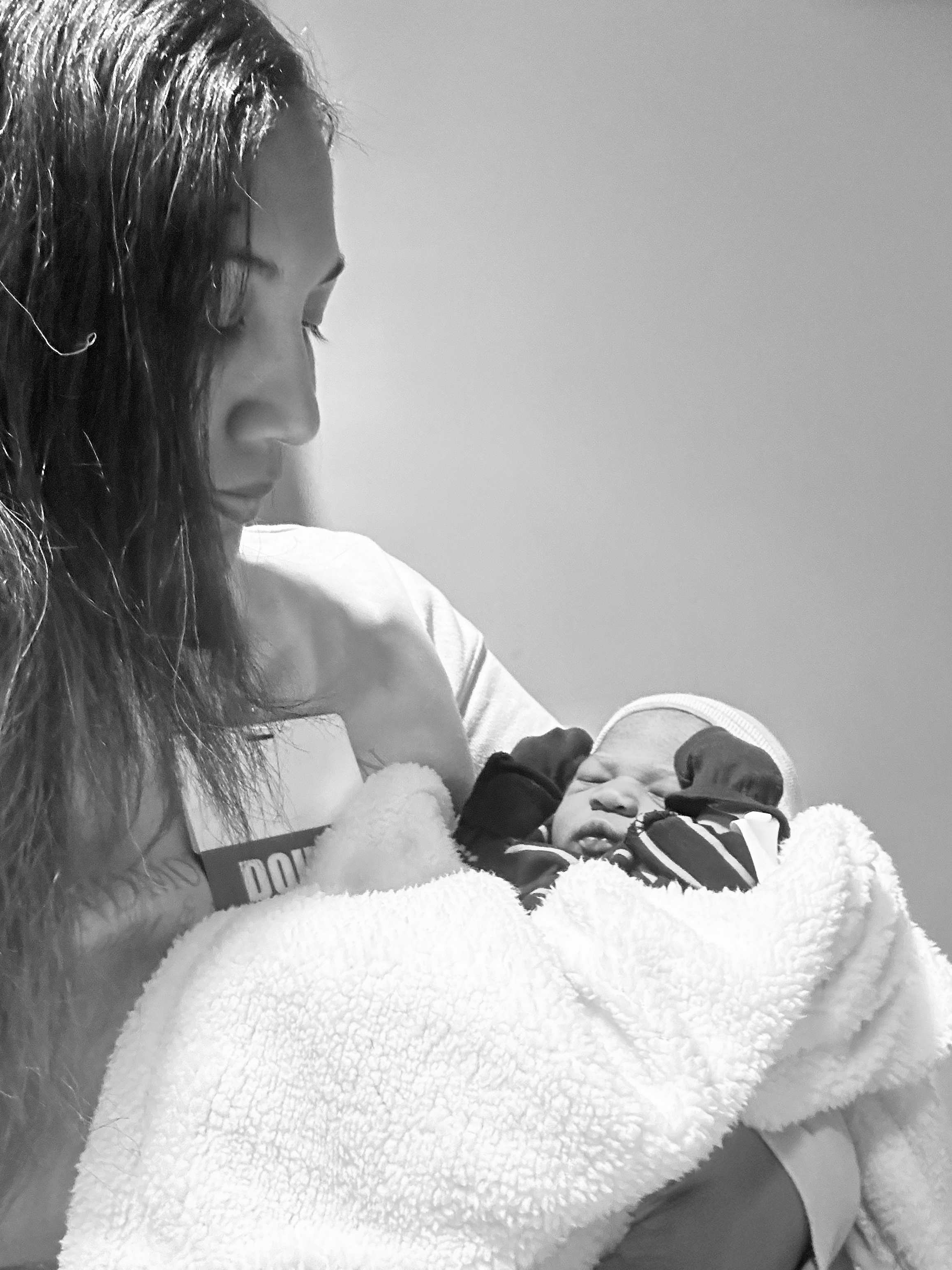 A woman with long hair and a wristband is sitting on a towel, holding and comforting a young child who is lying down on her lap, wearing a patterned jacket, while an emergency badge or patch is visible on her shirt.