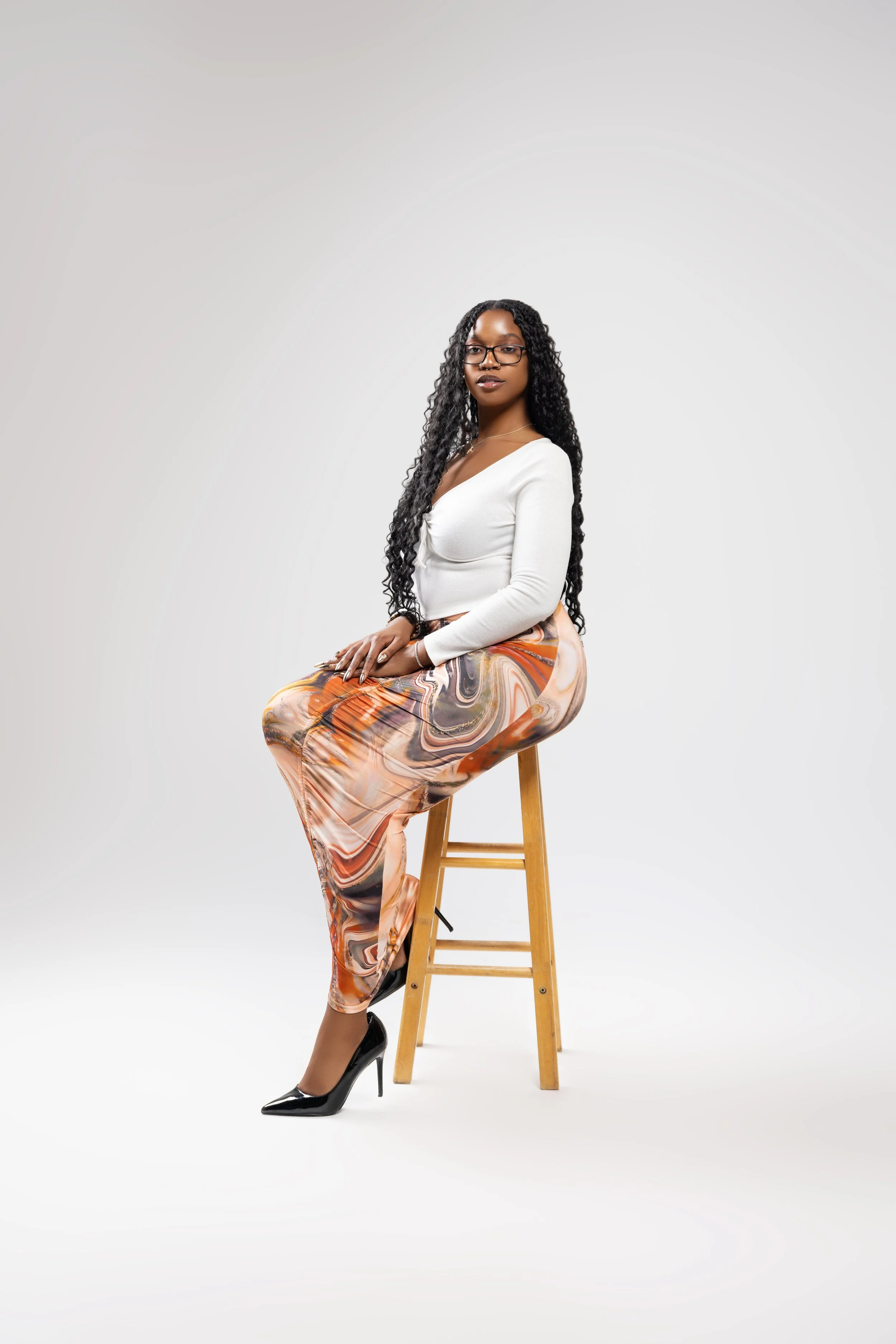 A woman with long, curly black hair, glasses, and dark lipstick sitting on a wooden stool, wearing a white long-sleeve top, colorful patterned pants, and black high heels against a plain white background.