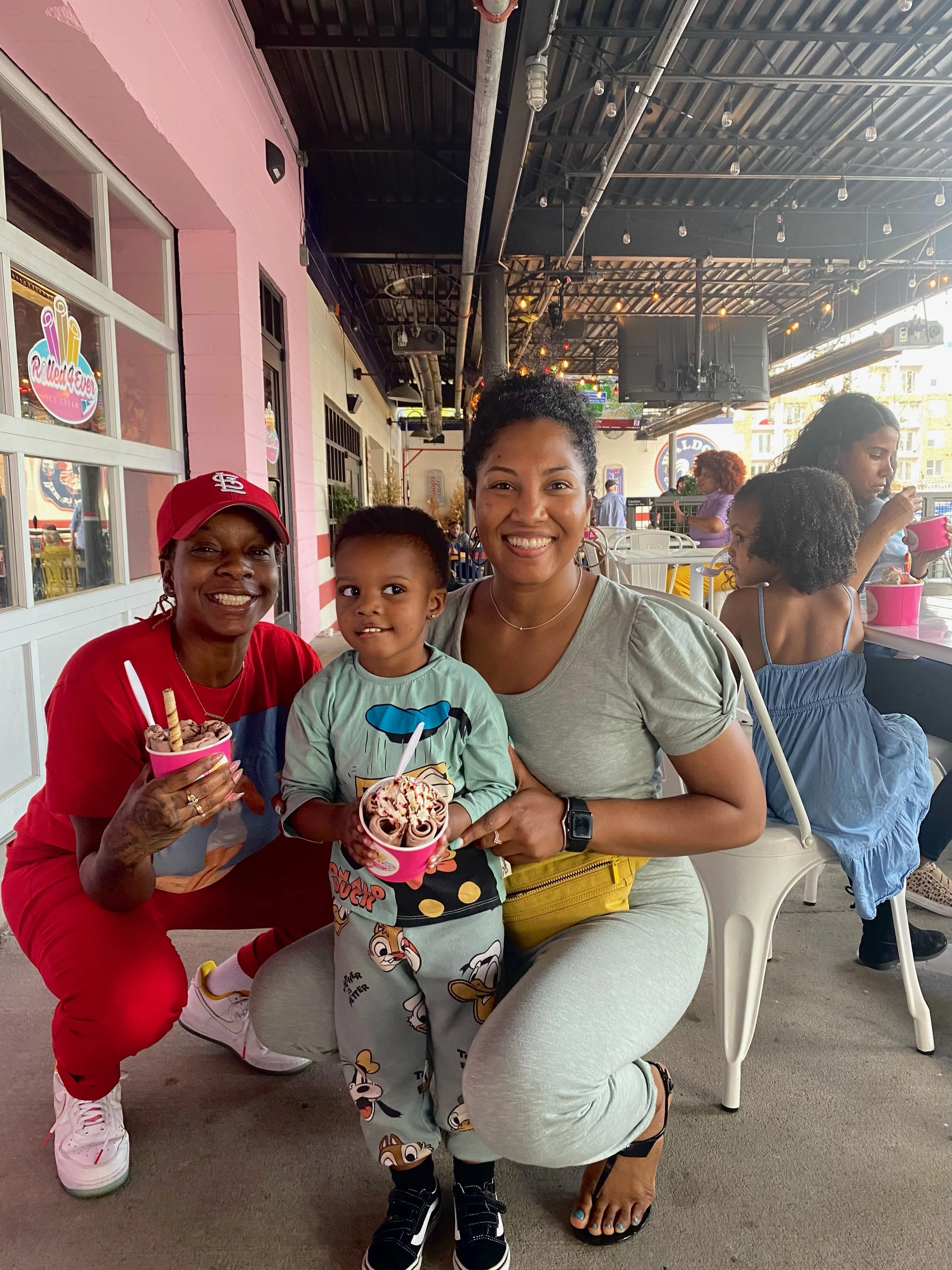 Three women and a young boy sitting at an outdoor ice cream shop, smiling and holding cups of ice cream. The woman on the left wears a red cap and red shirt, the woman in the middle wears a gray shirt, and the woman on the right wears a blue dress an