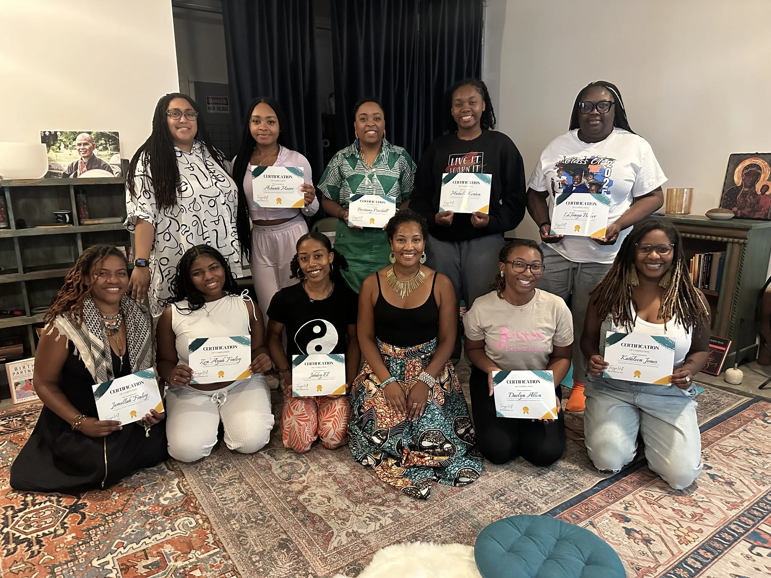 Group of women at an indoor event, some holding certificates, smiling for the photo. Decorations include framed art and books on shelves, with a patterned rug on the floor.
