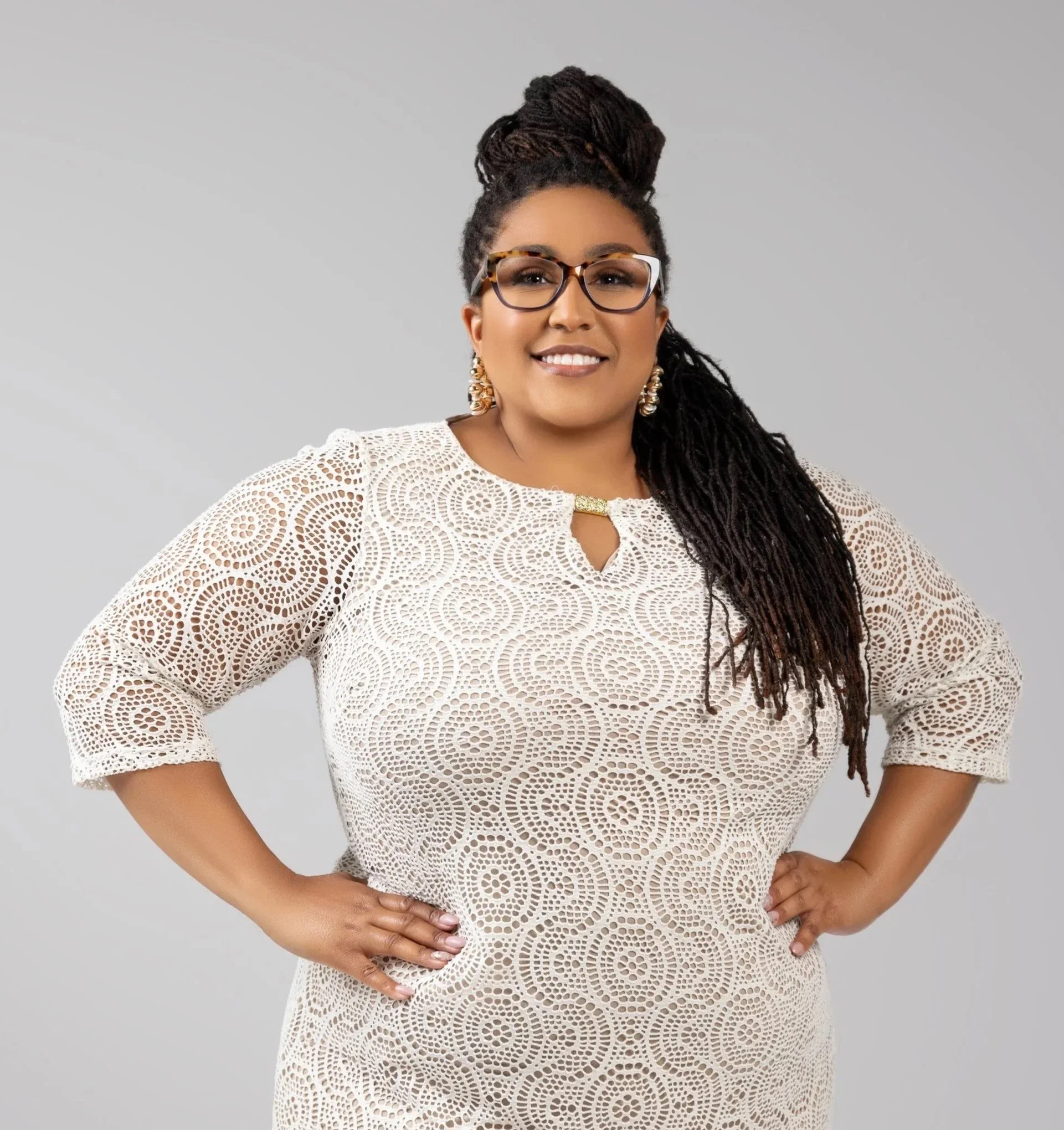 Woman with long braided hair wearing eyeglasses, gold earrings, and a white lace dress, standing against gray background.