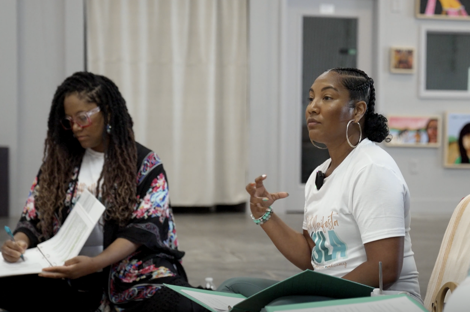 Two women are sitting at a table in a room decorated with pictures on the wall. The woman on the right is wearing a white t-shirt with teal and orange writing, large hoop earrings, and is gesturing with her hands. The woman on the left is wearing gla