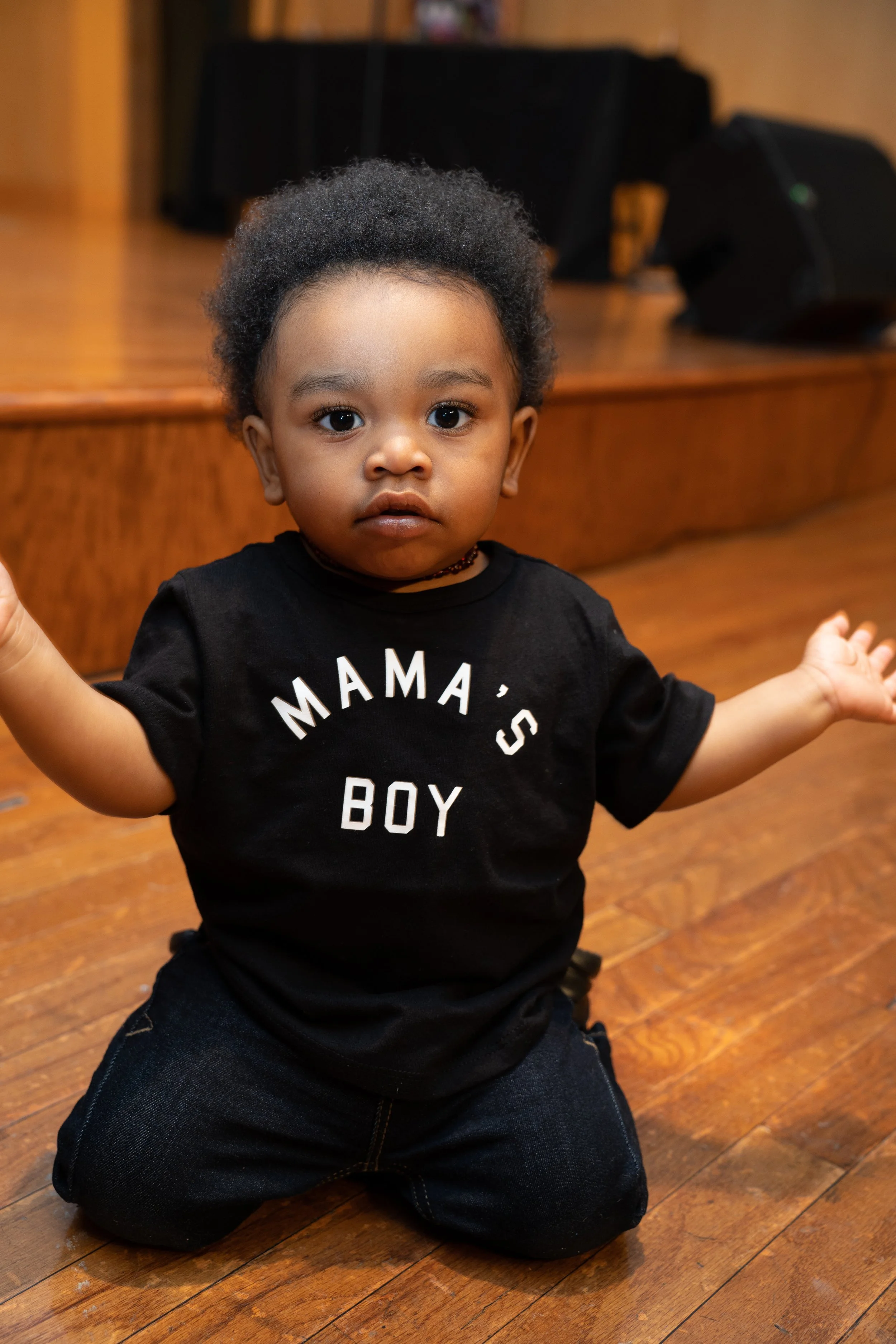 A young child with curly hair kneeling on a wooden floor with arms extended to the sides, wearing a black T-shirt that says 'MAMA'S BOY' and dark jeans, indoors with a blurred background.