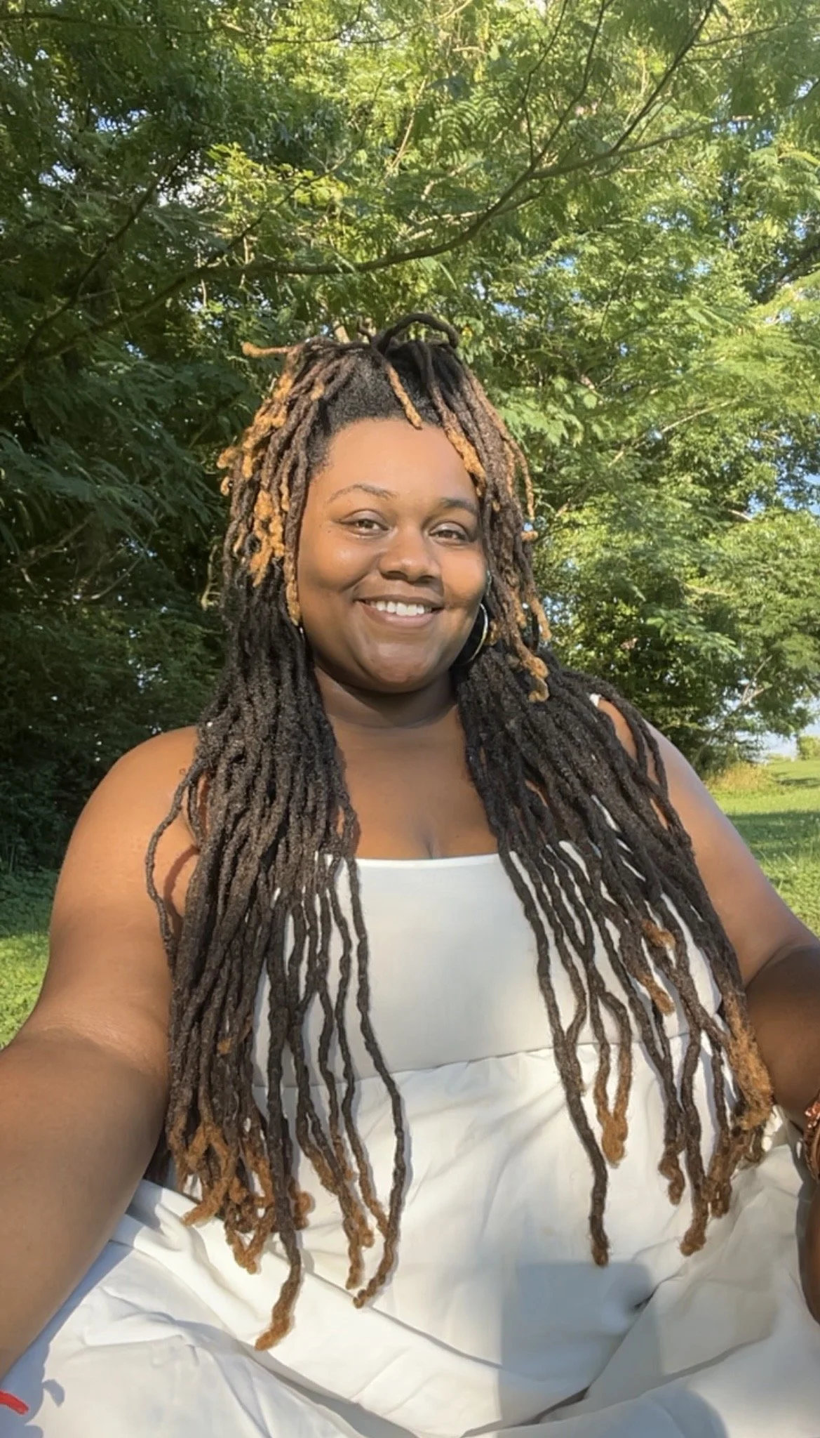 A smiling woman with long dreadlocks wearing a white top outdoors in a green, leafy area.