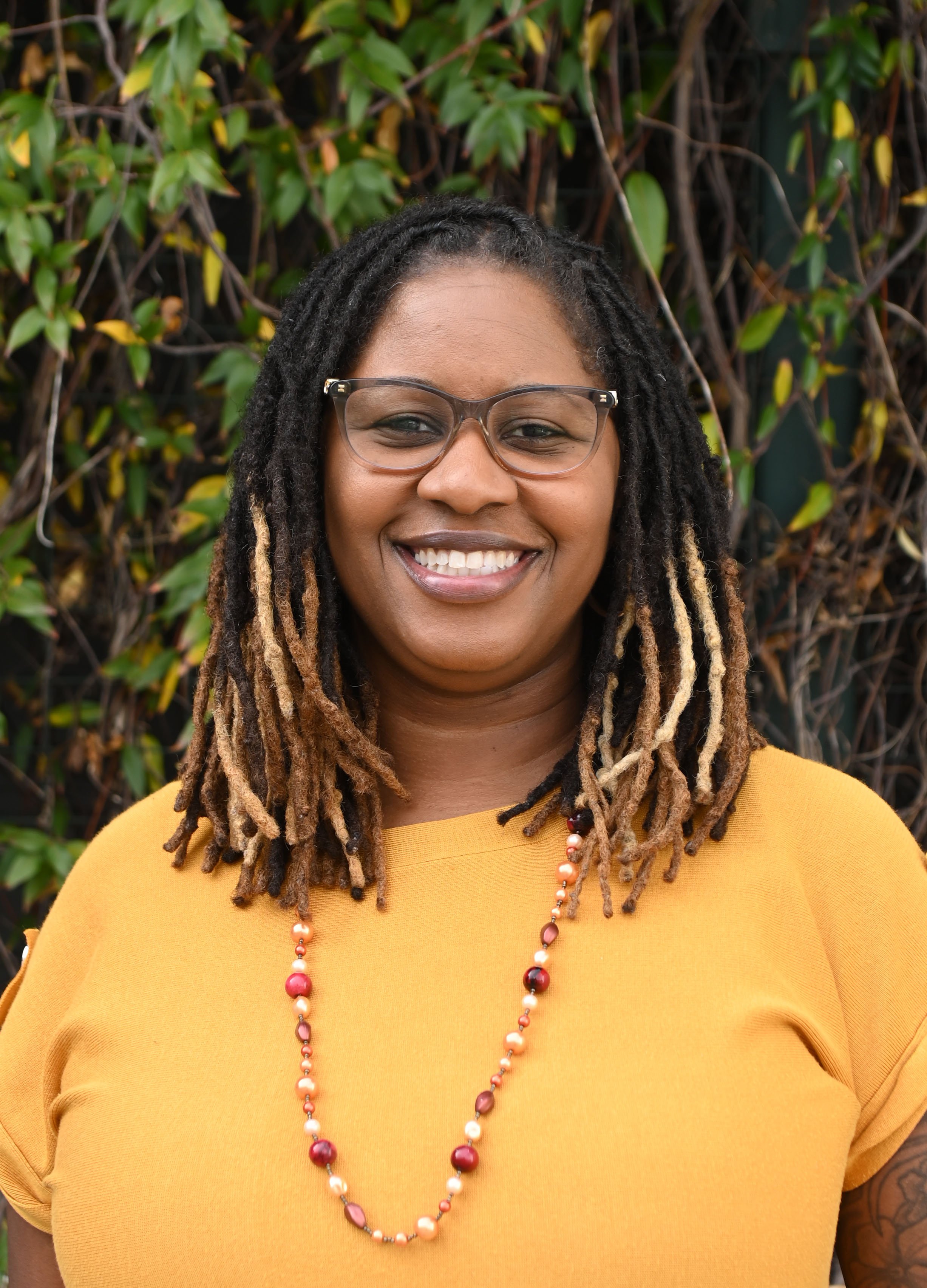 A woman with long dreadlocks and glasses, smiling, wearing a yellow top and a beaded necklace, standing outdoors in front of green leafy background.