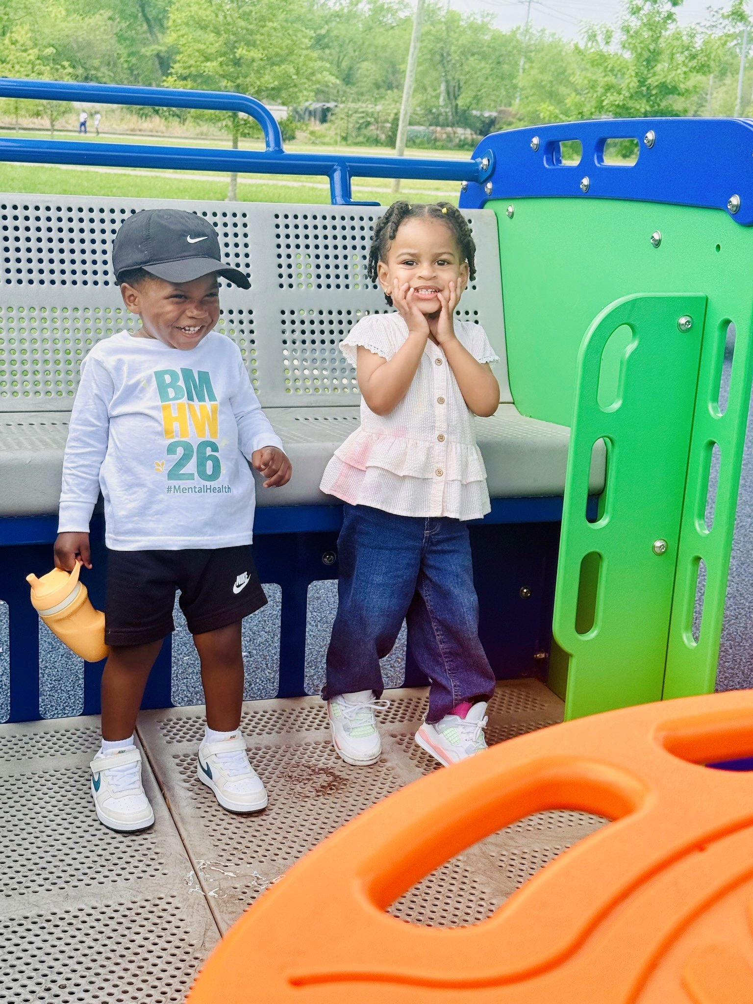 Two children, a girl and a boy, are standing on a playground structure, smiling and looking at the camera. The boy is wearing a black cap, a long sleeve white T-shirt with colorful text, black shorts, and white sneakers. The girl is wearing a light-c