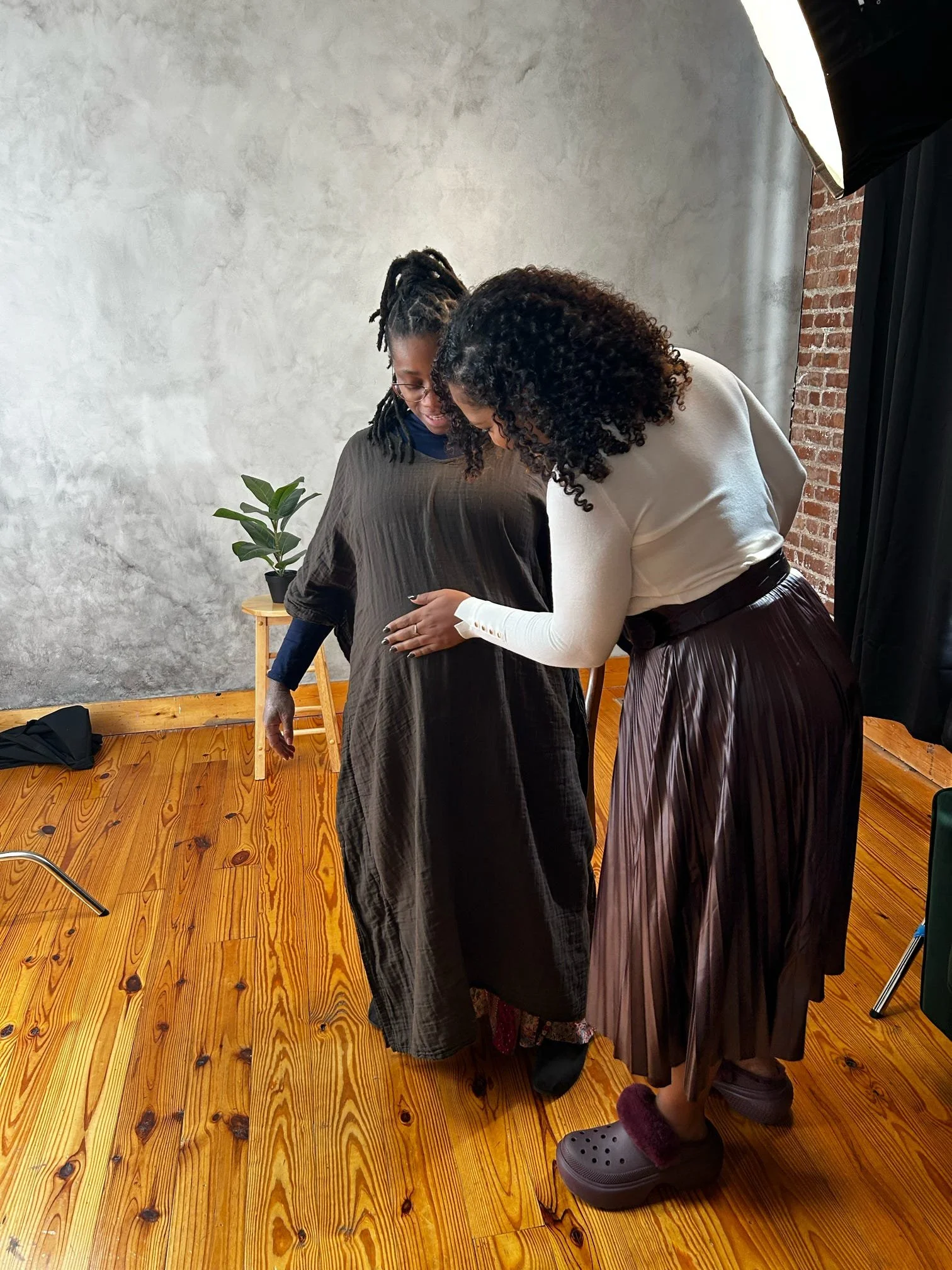 Two women are looking down at something, with one woman holding the other's waist. They are standing on a wooden floor in a room with a gray wall, a brick wall, and a small wooden table with a potted plant. One woman has dark curly hair, the other ha