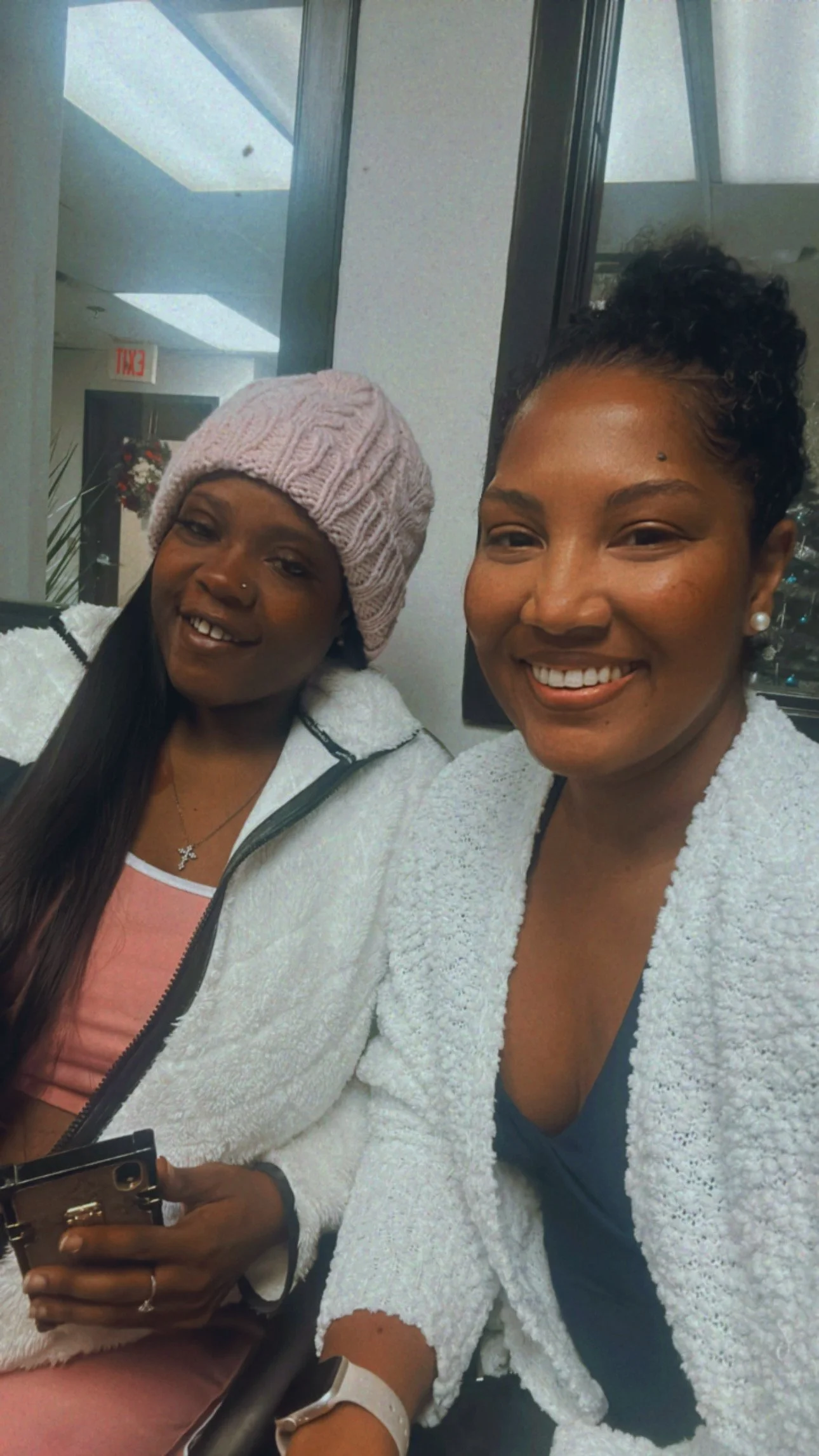 Two women taking a selfie together indoors, both smiling. One woman has short curly hair, wearing pearl earrings and a white cardigan. The other woman is wearing a pink knit hat, long straight hair, and a white zip-up jacket. The background includes 