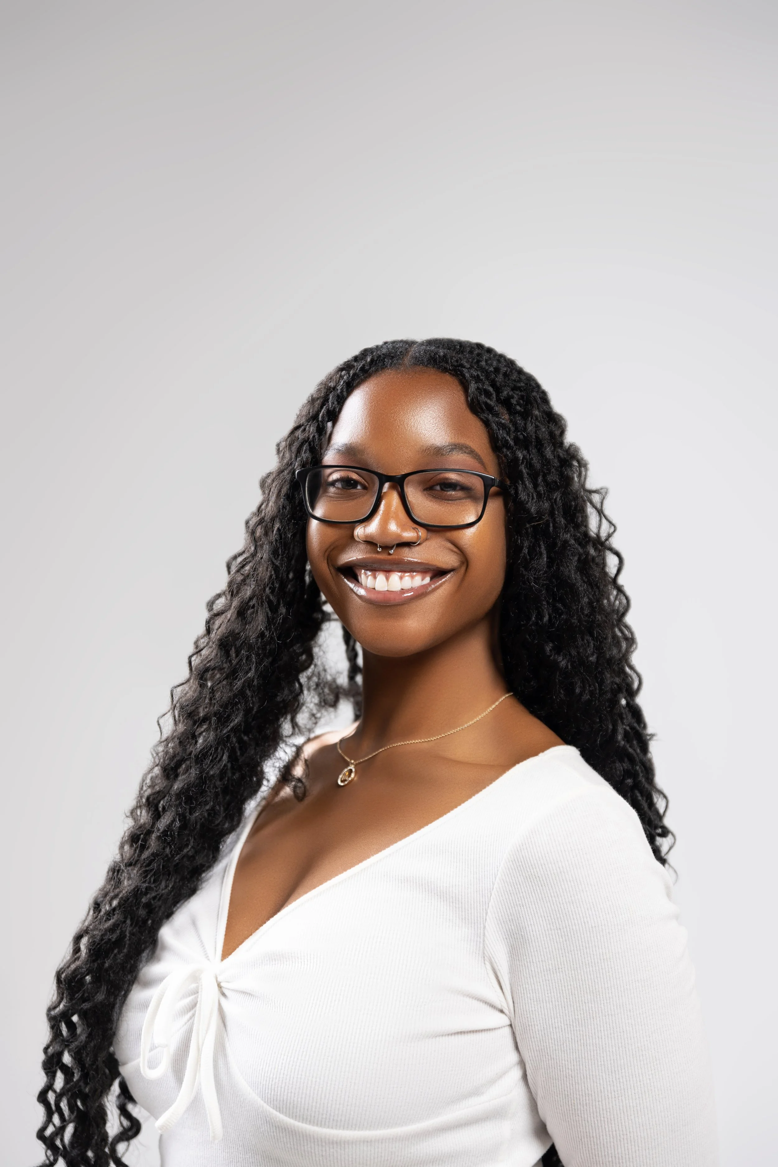 A smiling Black woman with long curly hair, wearing glasses, a nose ring, a necklace, and a white top with a tie-front detail, against a plain light gray background.