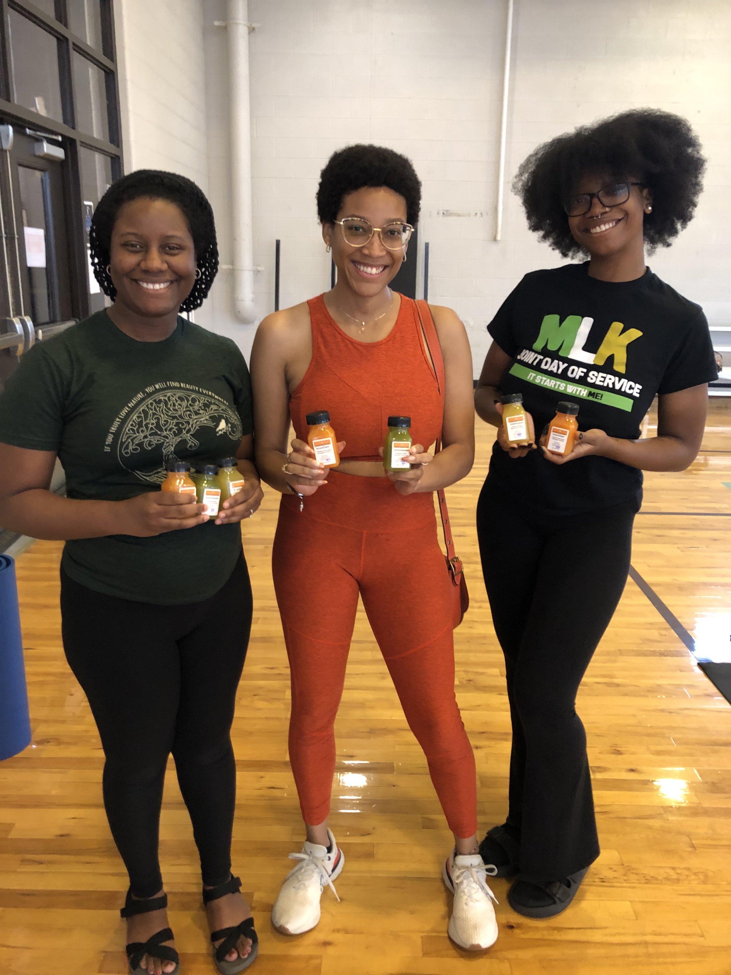 Three women standing indoors on a wooden floor, holding small bottles, smiling at the camera. The woman on the left wears a black T-shirt with gold design, the woman in the center wears an orange sports outfit with glasses, and the woman on the right