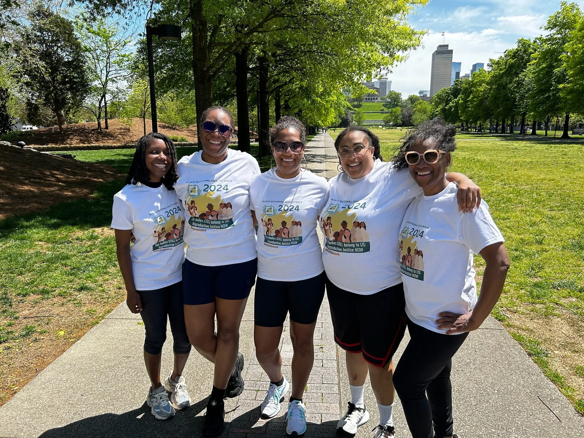 Five women standing together outdoors on a sunny day, smiling. They are wearing white t-shirts with a logo for the Black Maternal Health Walk 2024, and they are in a park with trees and a city skyline in the background.