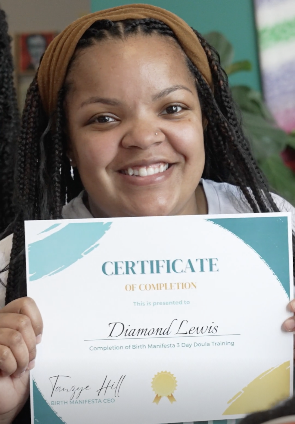 A woman with braided hair, wearing a brown headband, holding a certificate and smiling at the camera.