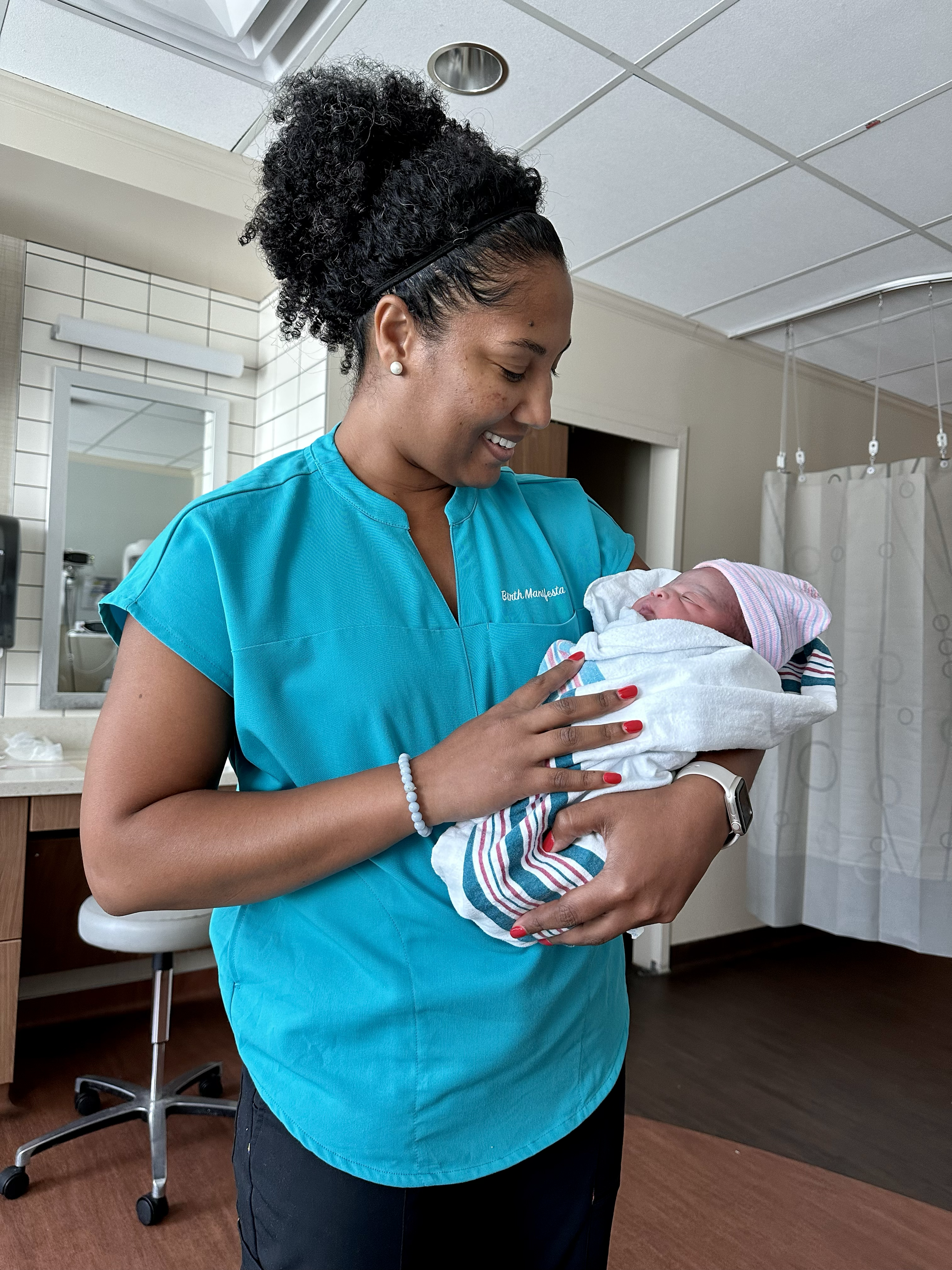 A woman in scrubs holding a newborn baby wrapped in a hospital blanket, smiling and looking at the baby, in a hospital room.