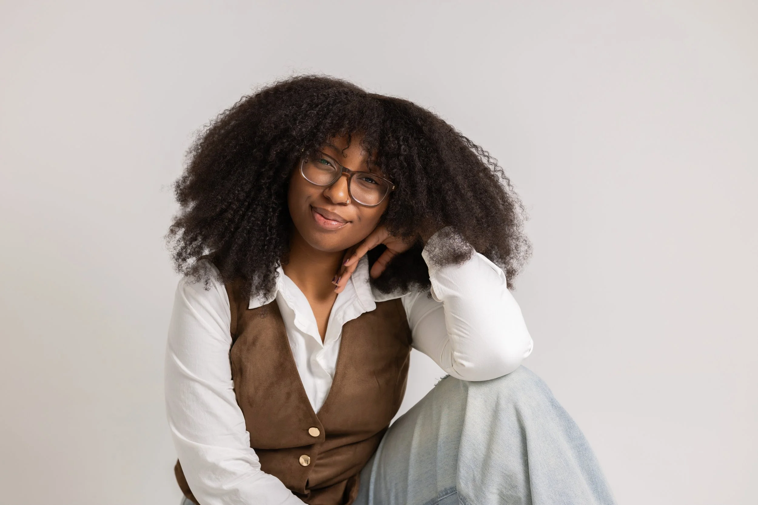 A woman with curly hair and glasses posing against a plain white background, resting her hand on her face and smiling.