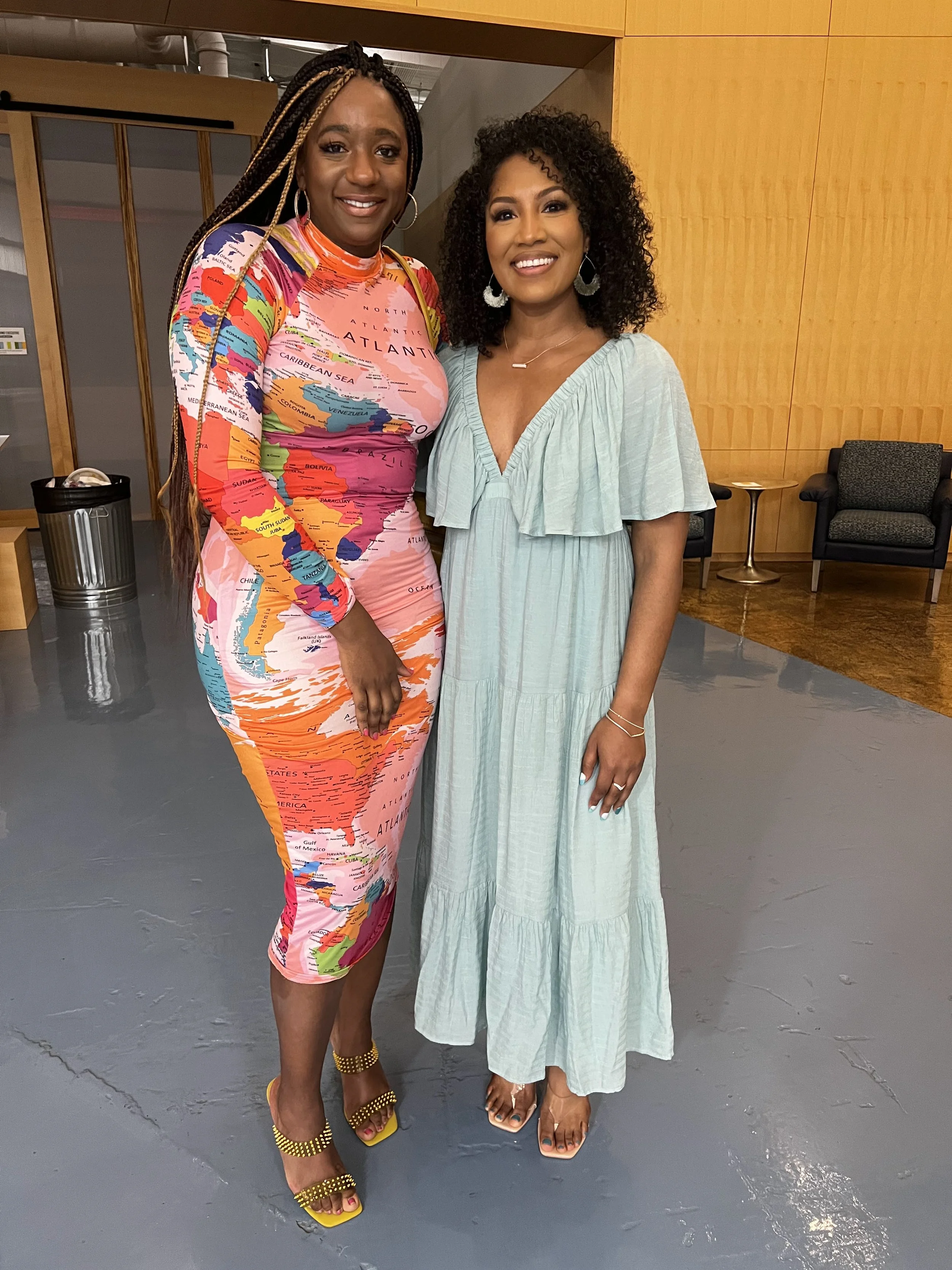 Two women standing together in a room with yellow wood paneled wall, smiling at the camera. One woman wears a colorful dress with a world map print, and the other wears a light green dress. They are wearing earrings and sandals.