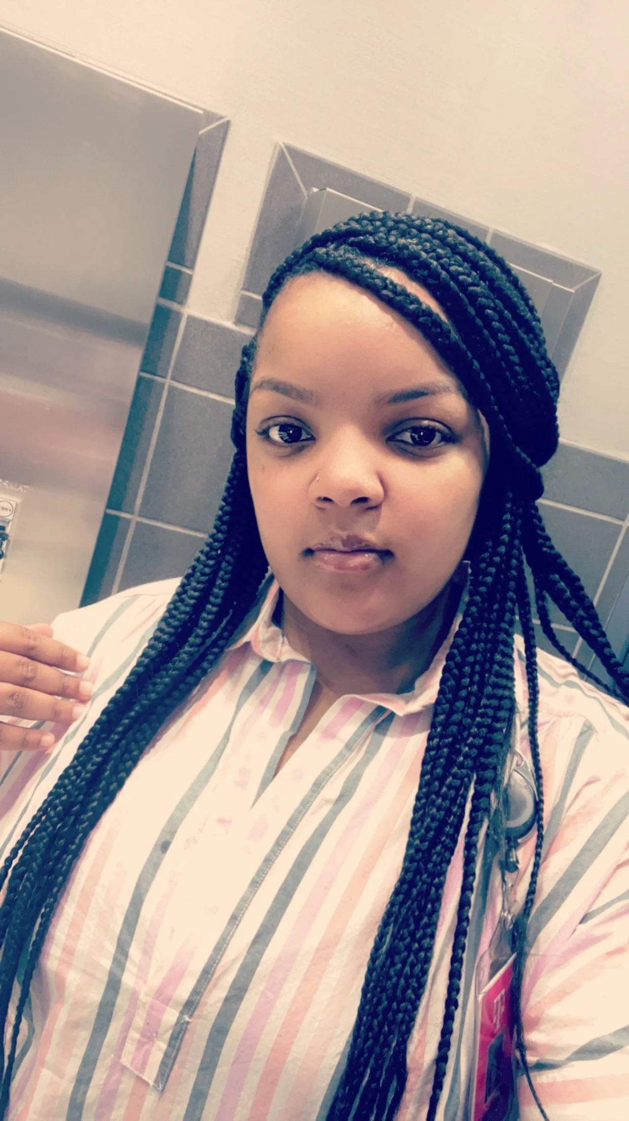 A woman with long braided hair wearing a striped shirt in a bathroom with gray tiles and a mirror.