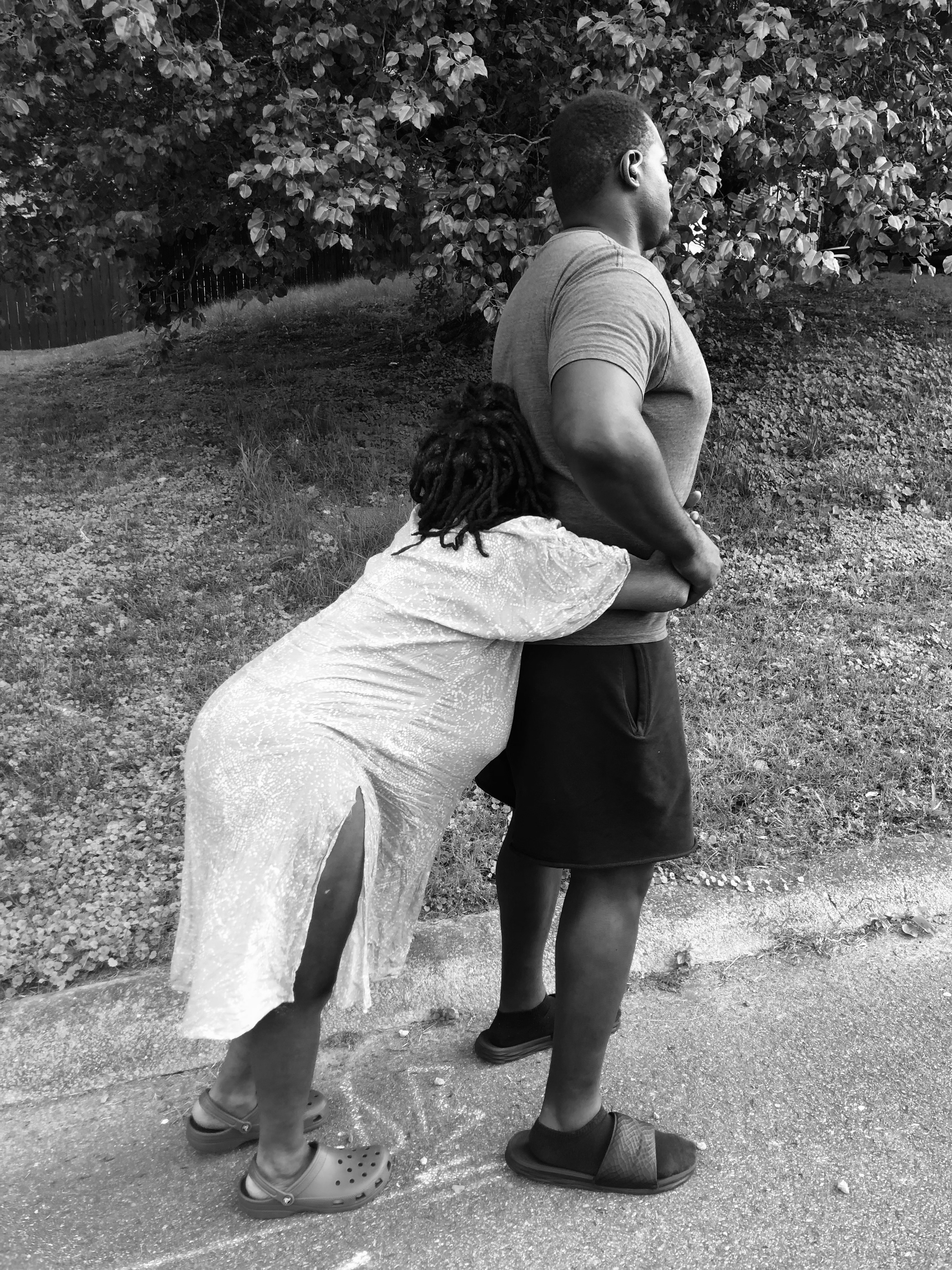 A person with short hair is standing outdoors with their hands on their hips, wearing a t-shirt and shorts. A woman with long dreadlocks, wearing a light-colored dress, is hugging their leg from behind. Both are wearing slip-on shoes, and there are t
