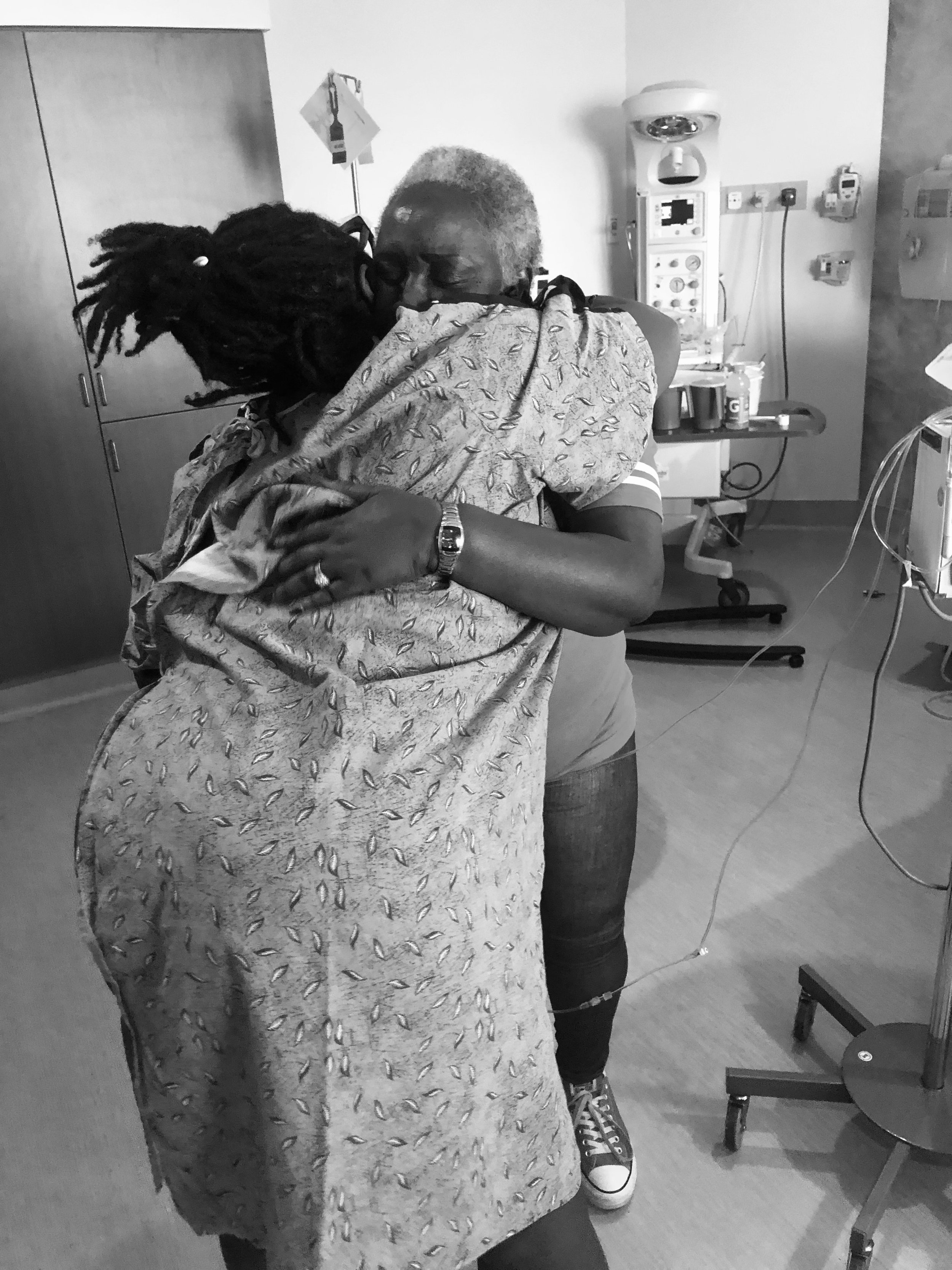 A person with gray hair and a colorful shirt hugging a person with dreadlocks, wearing a patterned dress in a medical setting, with hospital equipment and furniture around.