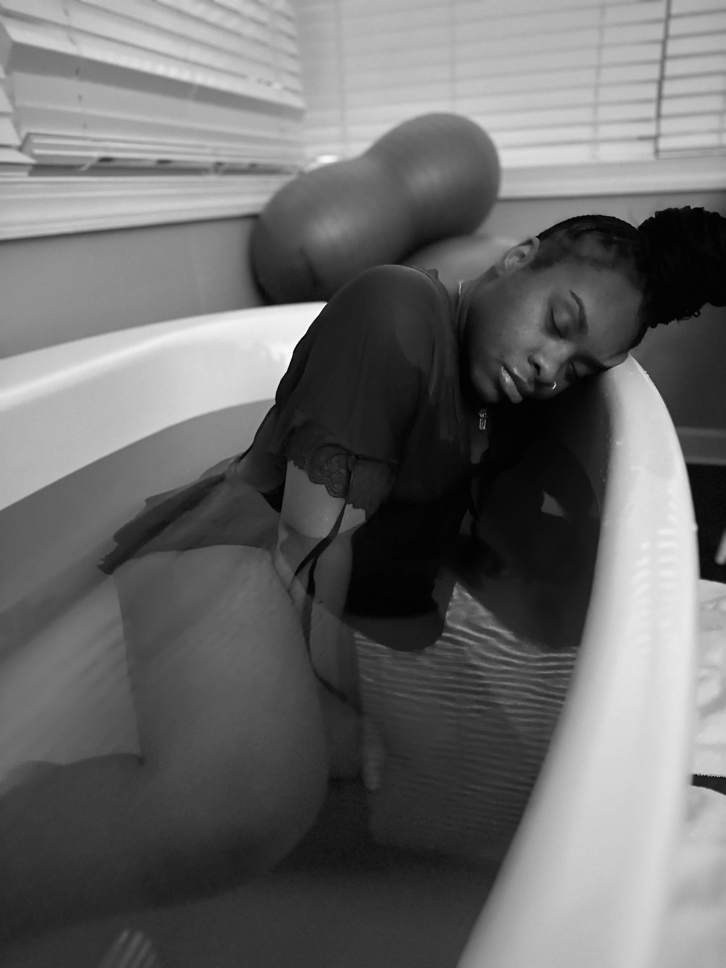 A woman with braids and a nose ring resting her head on her crossed arms, sitting in a bathtub in a bathroom near closed window blinds.
