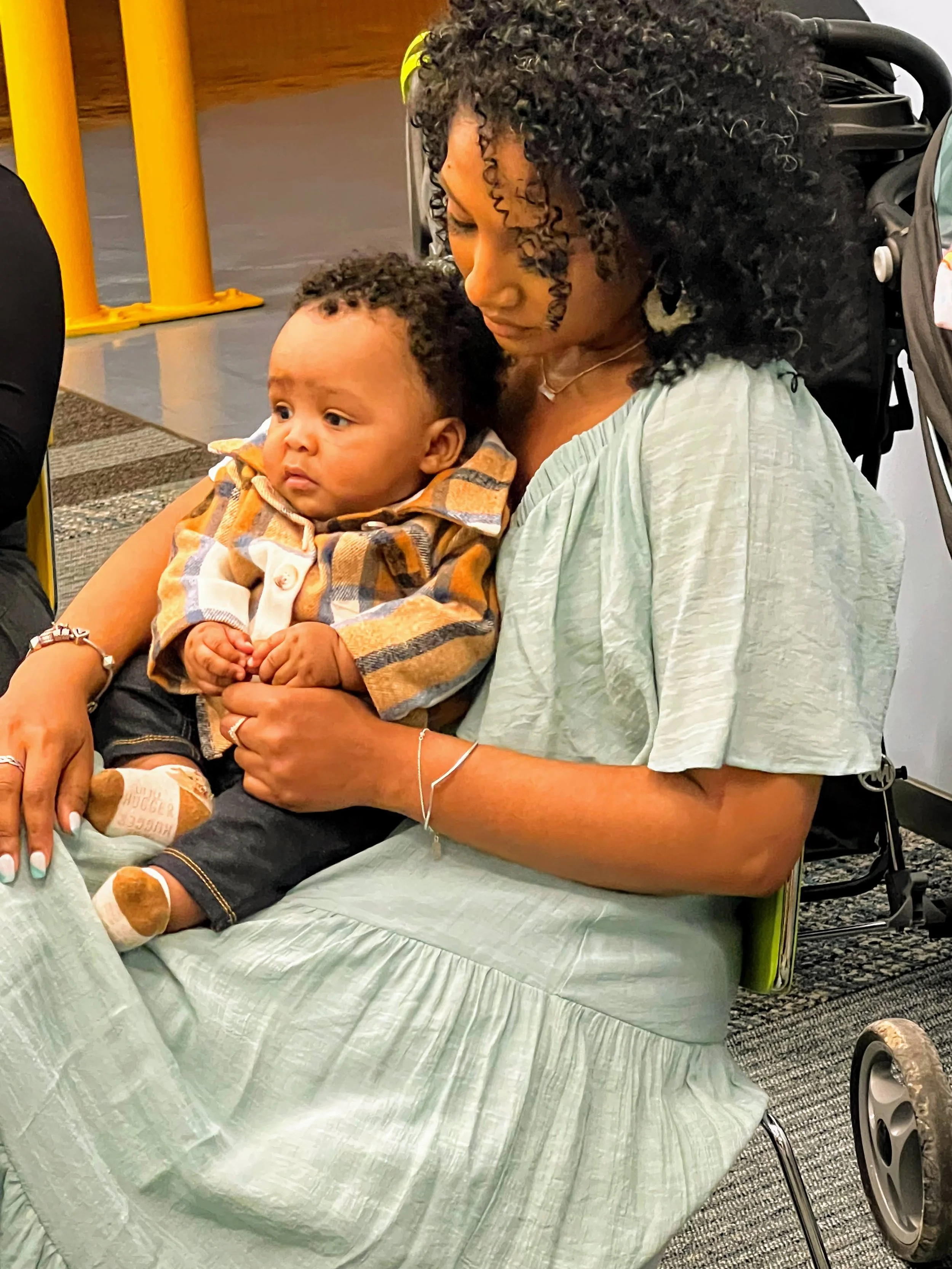 A woman with curly black hair holding a young child with curly hair sitting on her lap. They are sitting on a hospital bed or chair with a green sheet, and the woman appears to be in a hospital or medical facility.