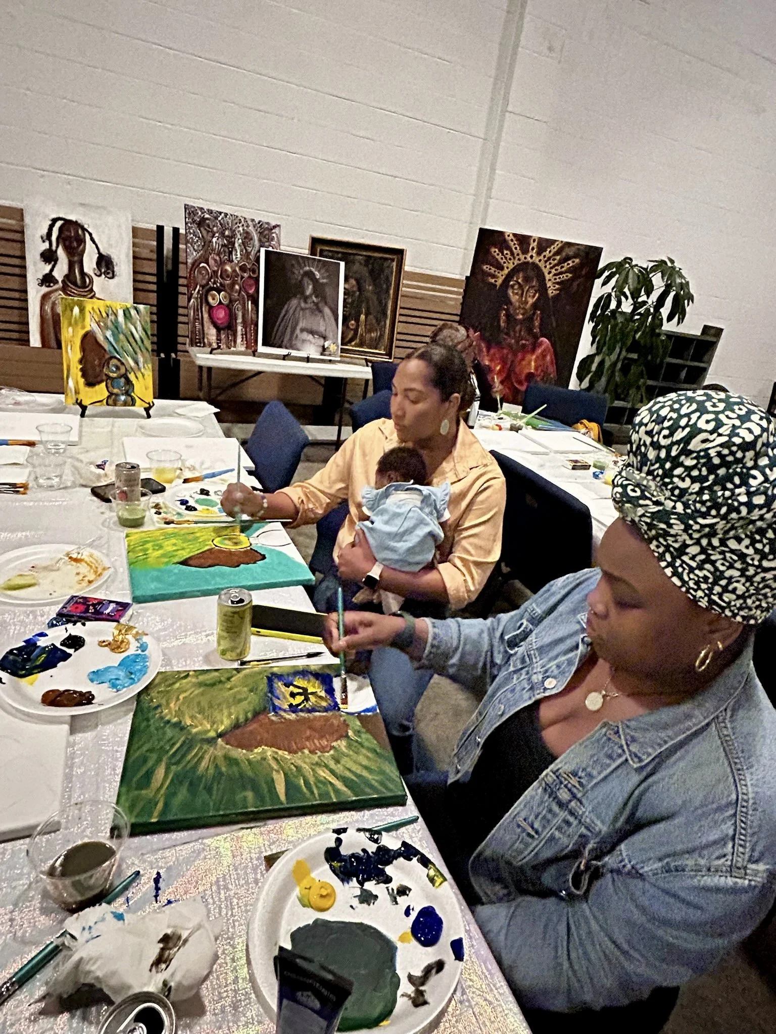 People painting canvases at an art event with paintings of African women and religious figures displayed on tables and stands, and a large houseplant in the background.