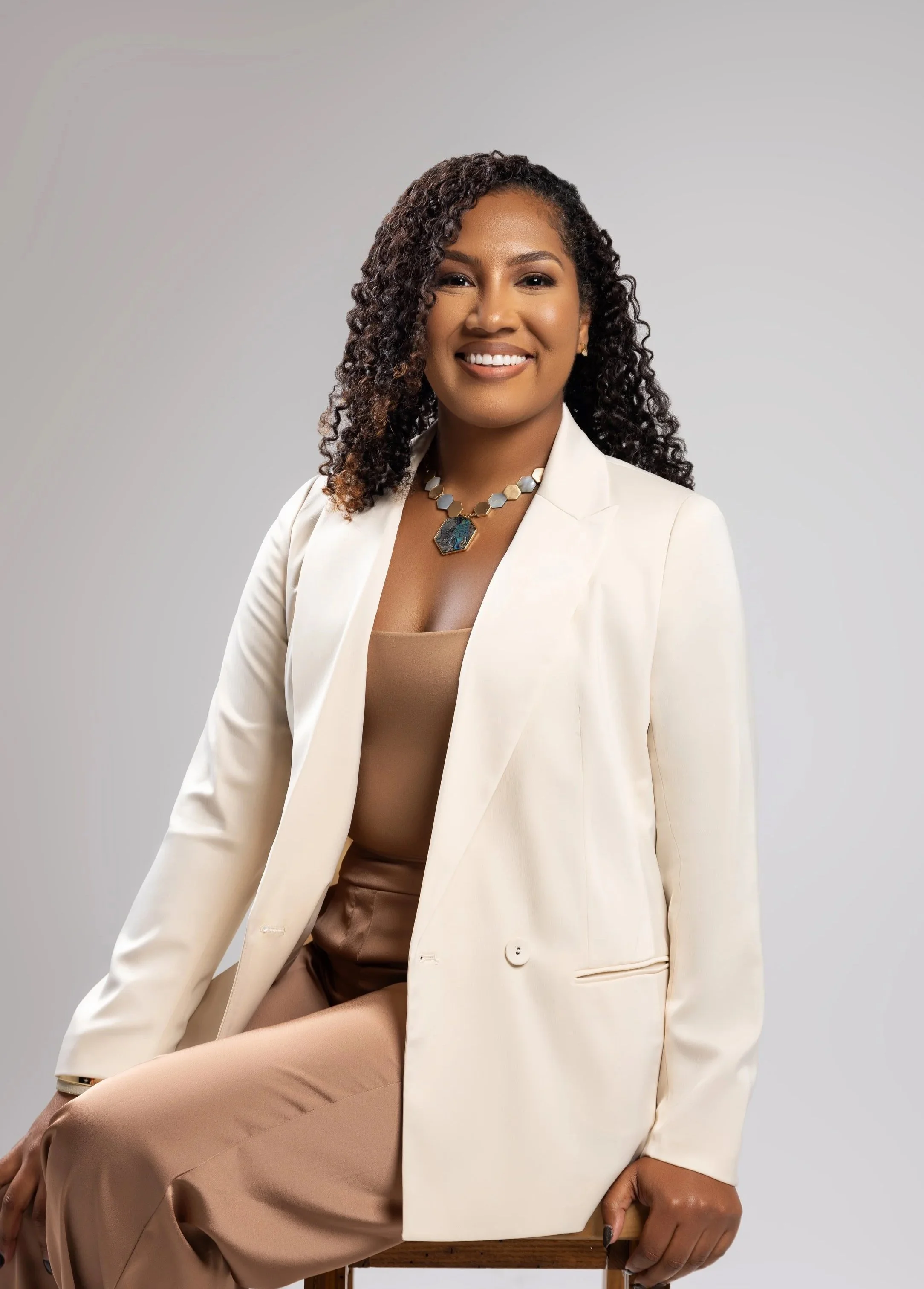 A smiling woman with curly dark hair wearing a cream blazer, beige top, and brown pants, sitting on a wooden chair against a plain light gray background.