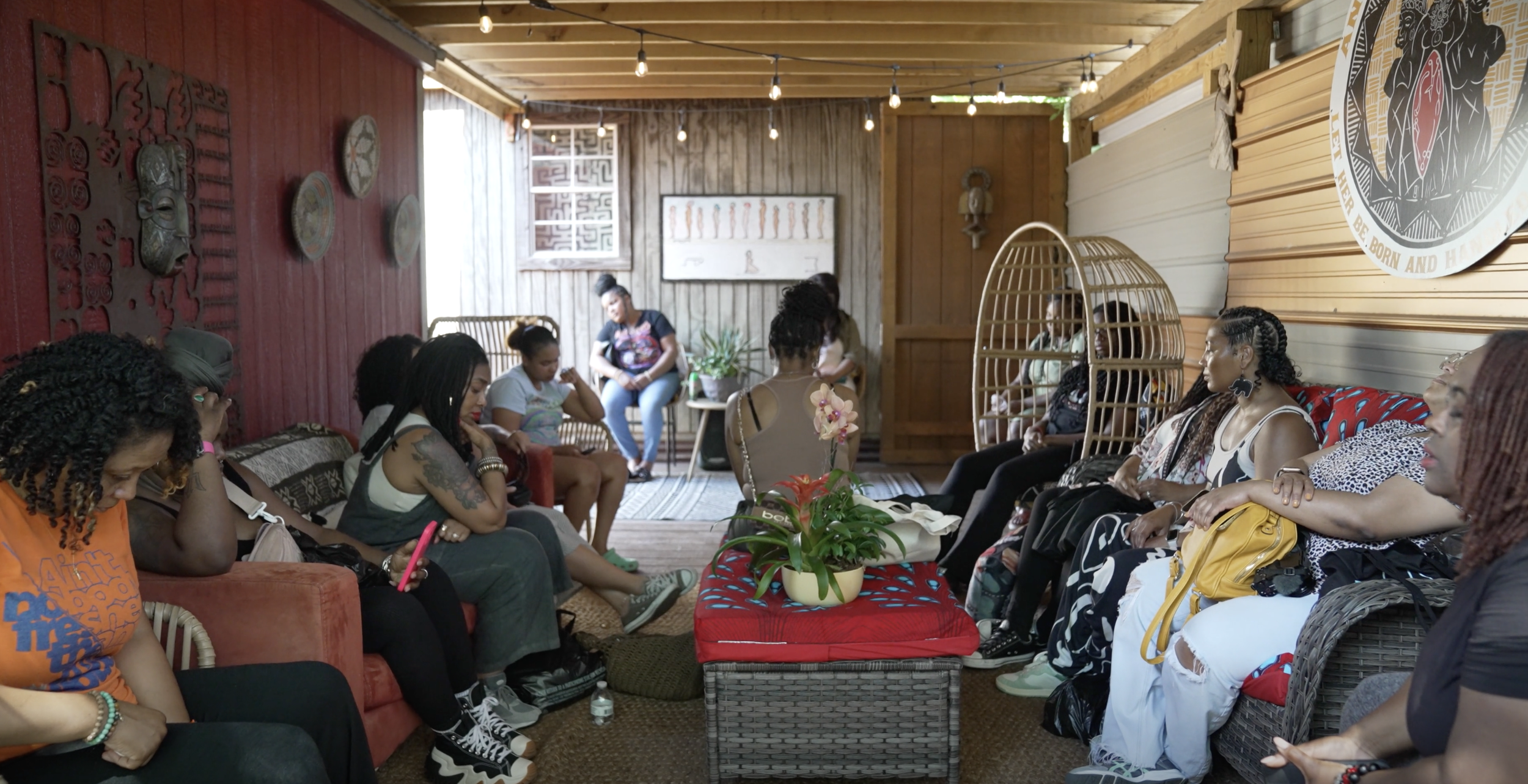 A group of women sitting in a cozy indoor space with wooden walls and ceiling, some in hanging chairs, during a gathering or meeting, with decorative art and plants.