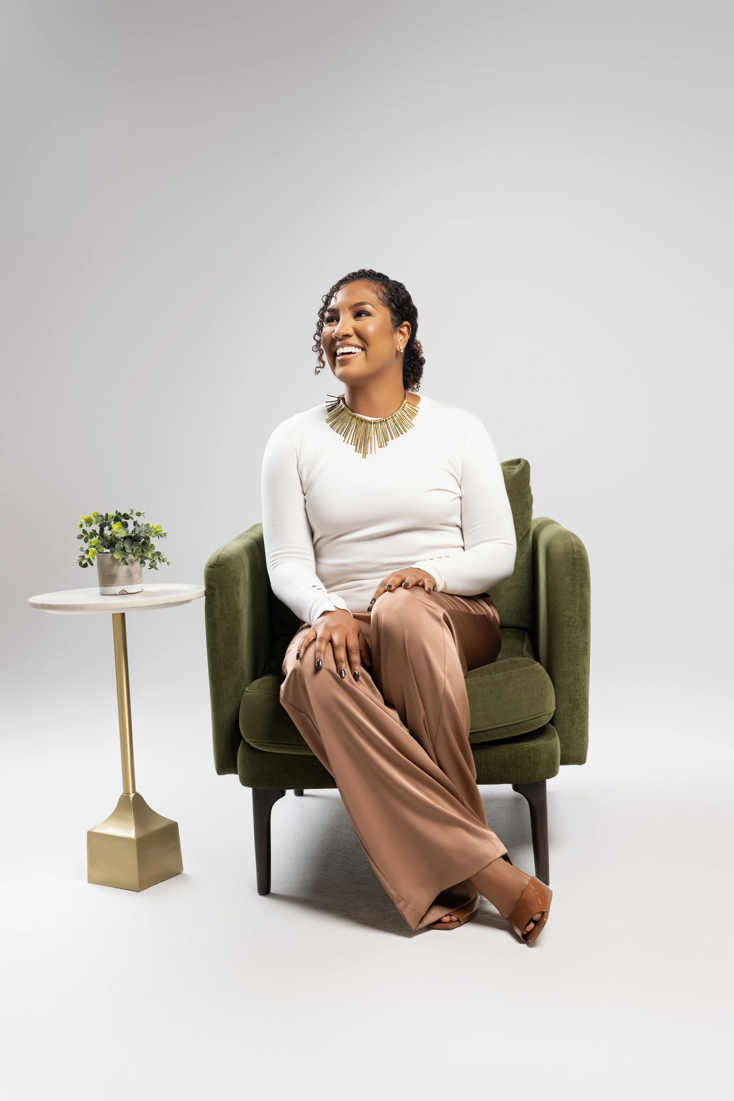 A woman with short curly hair sitting on an olive green armchair, smiling, wearing a white long sleeve top, beige pants, a gold statement necklace, and brown open-toe shoes. There is a small round side table with a beige vase and green plant to her left.