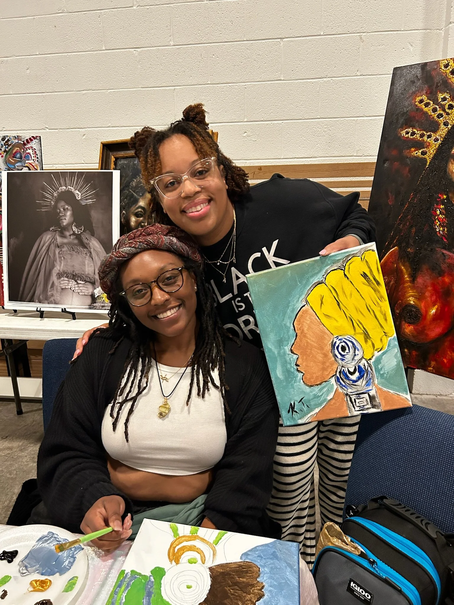 Two women are at an art table displaying completed paintings and art supplies. The woman in front has dreadlocks and glasses, smiling at the camera, holding a paintbrush. The woman behind has hair styled in two buns, wearing glasses, and is also smil