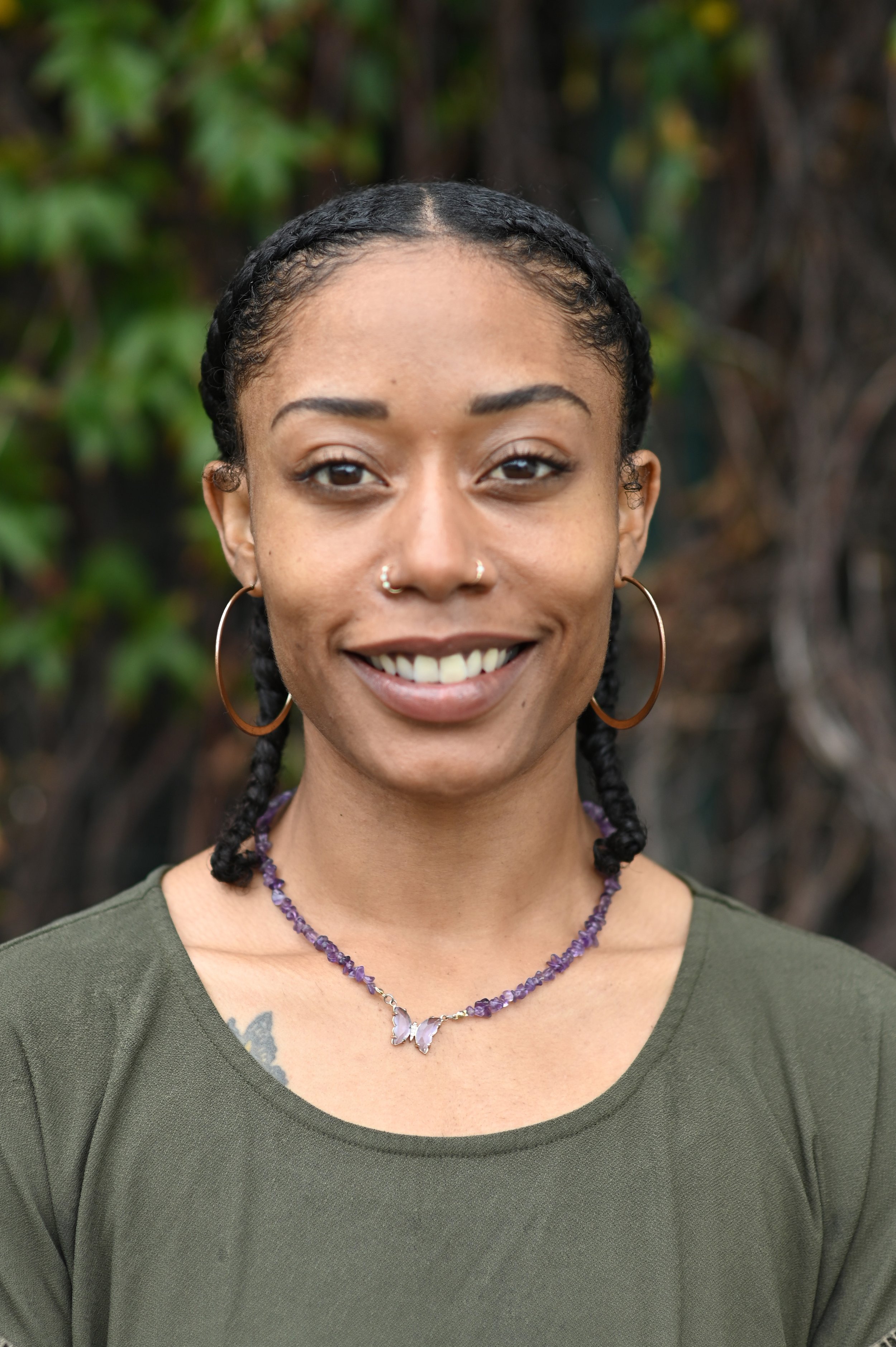 Close-up portrait of a smiling woman with braided black hair, hoop earrings, and a purple beaded necklace, wearing a green shirt, outdoors with blurred green foliage in the background.