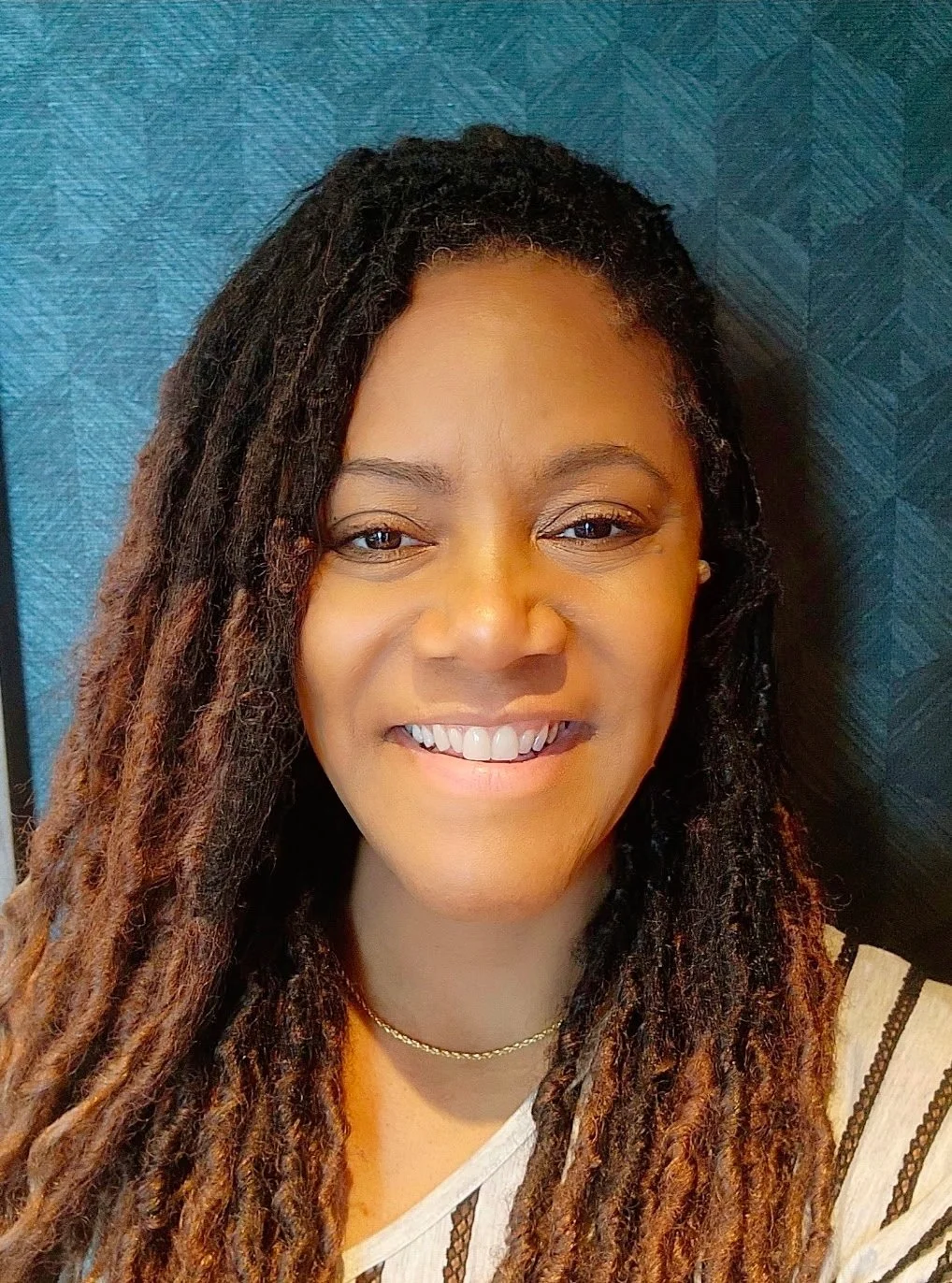 Close-up of a smiling woman with long dark dreadlocks, wearing a striped top and a gold necklace, standing in front of a textured blue background.