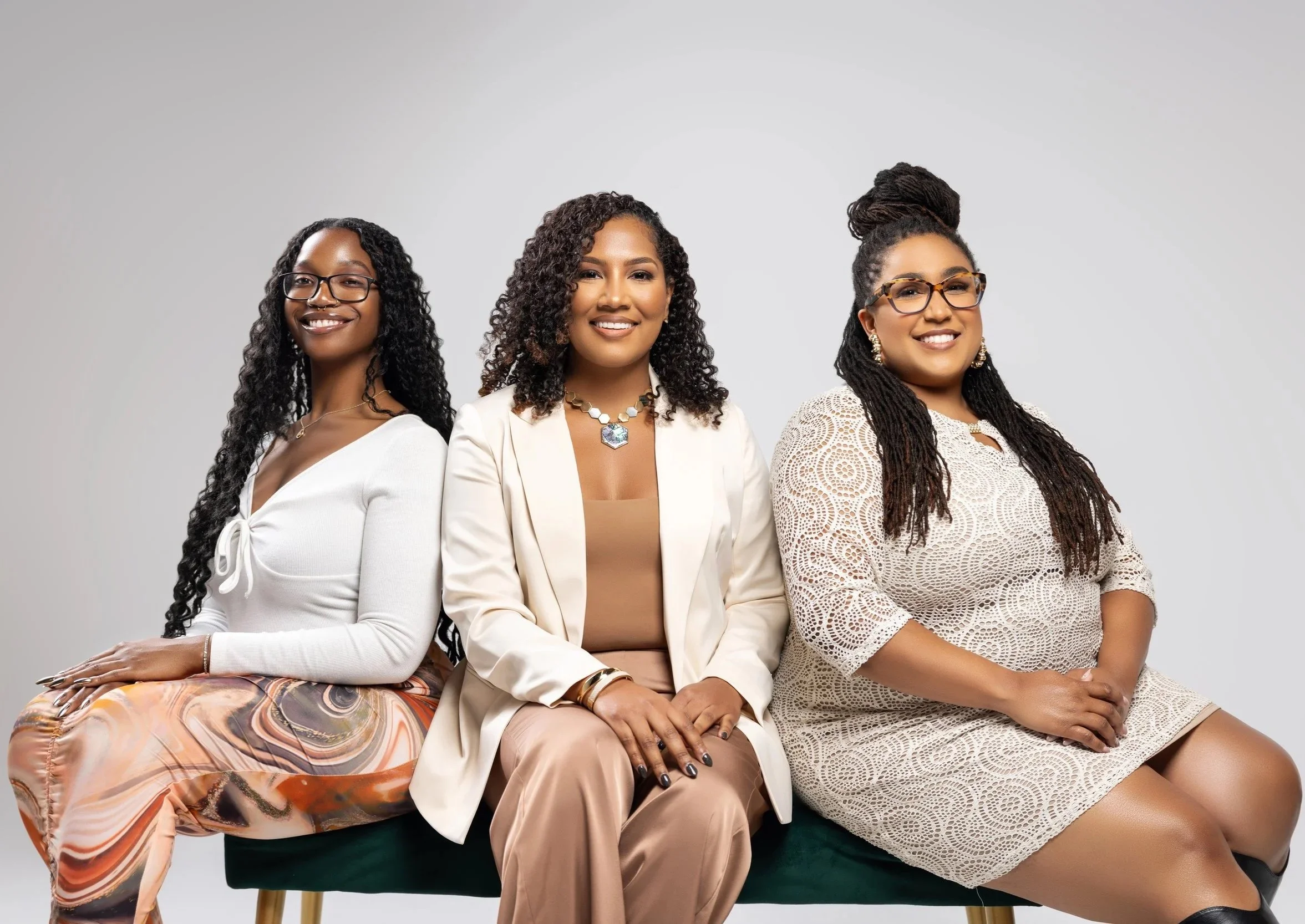 Three women sitting together against a plain gray background. They are smiling and dressed stylishly with glasses and jewelry.