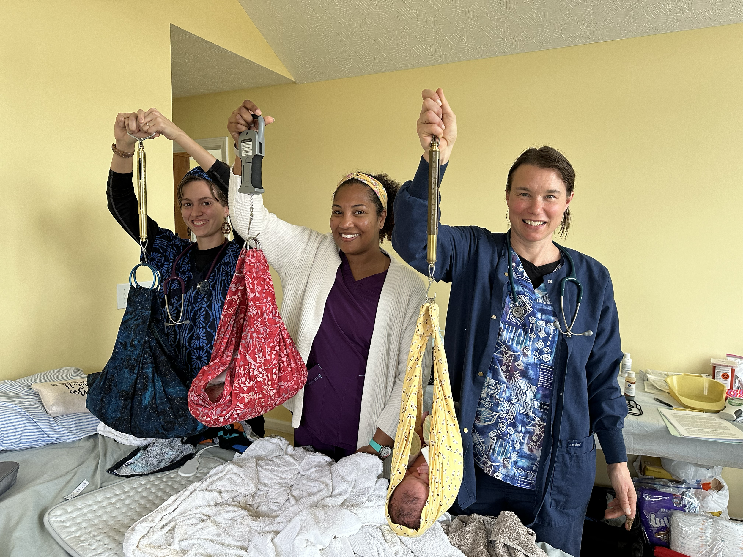 Three healthcare workers, including two women, hold medical instruments and colorful fabric bags while standing in a hospital room with a bed and medical supplies. They are smiling and appear to be celebrating a successful delivery.