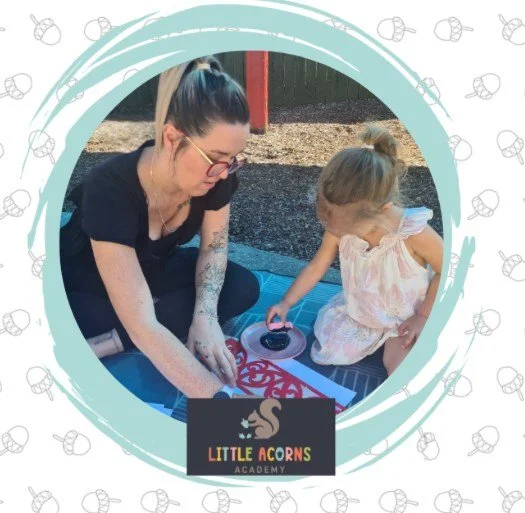 A woman and a young girl are sitting on a blue mat outdoors. The woman is wearing glasses, has dark hair tied back, and is dressed in black. The girl, wearing a light pink dress, is playing with a small toy. The background includes a playground structure and dirt ground. There is a logo for 'Little Acorns Academy' at the bottom of the image.