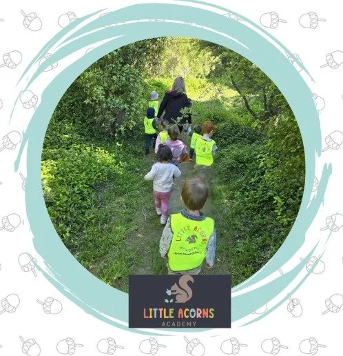 Children and an adult walking on a nature trail surrounded by greenery, with some children wearing yellow reflective vests that say 'Little Acorns Academy'.