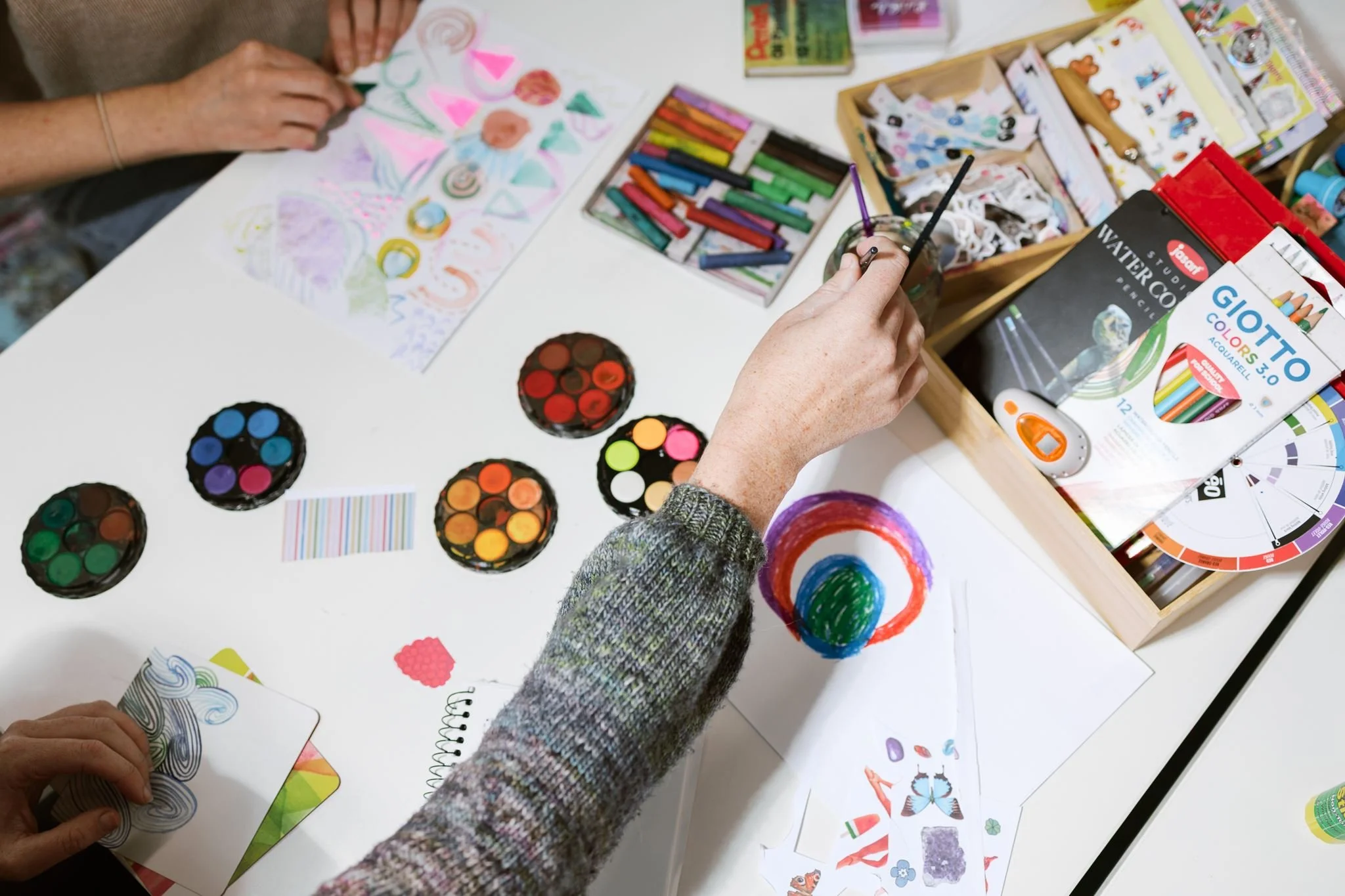 Hands of people creating colorful artwork with markers, paint, and drawing supplies on a white table, surrounded by art materials and colorful sketches.